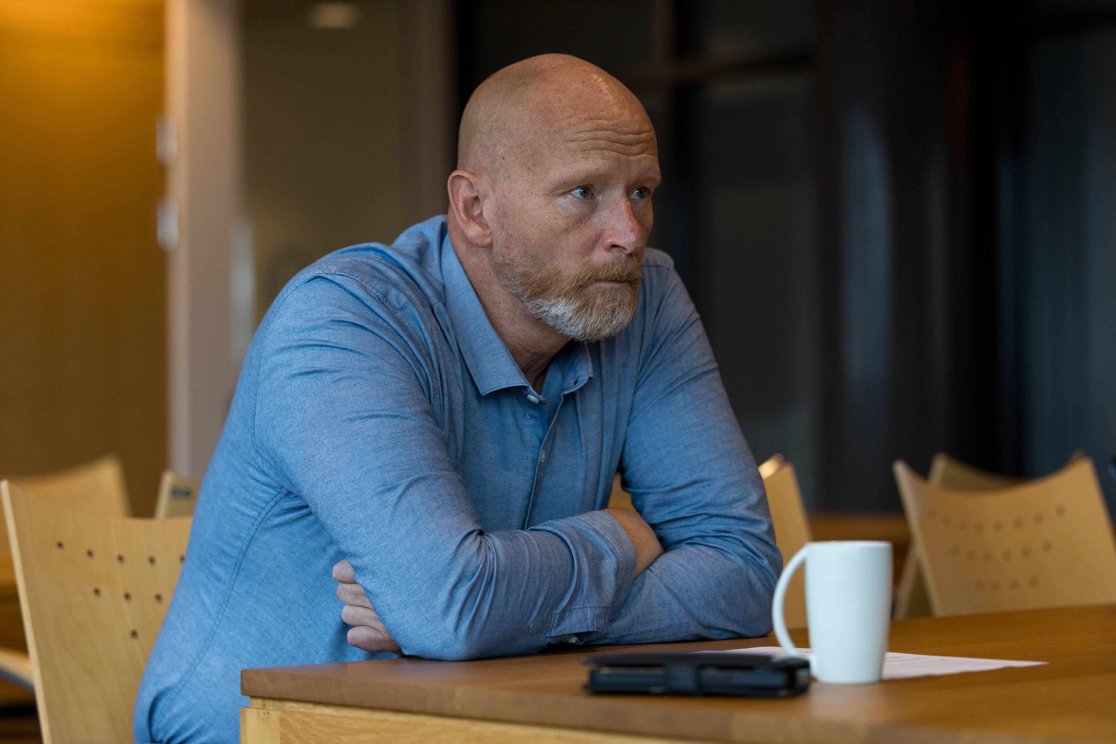 Svindeljeger i Sparebank 1, Erik Bjerkaas, sitter rundt et bord og lener seg på albuene. Han har lyseblå skjorte, skjegg, og ser alvorlig ut. Foran ham på bordet er en hvit kaffekopp og mobilen.