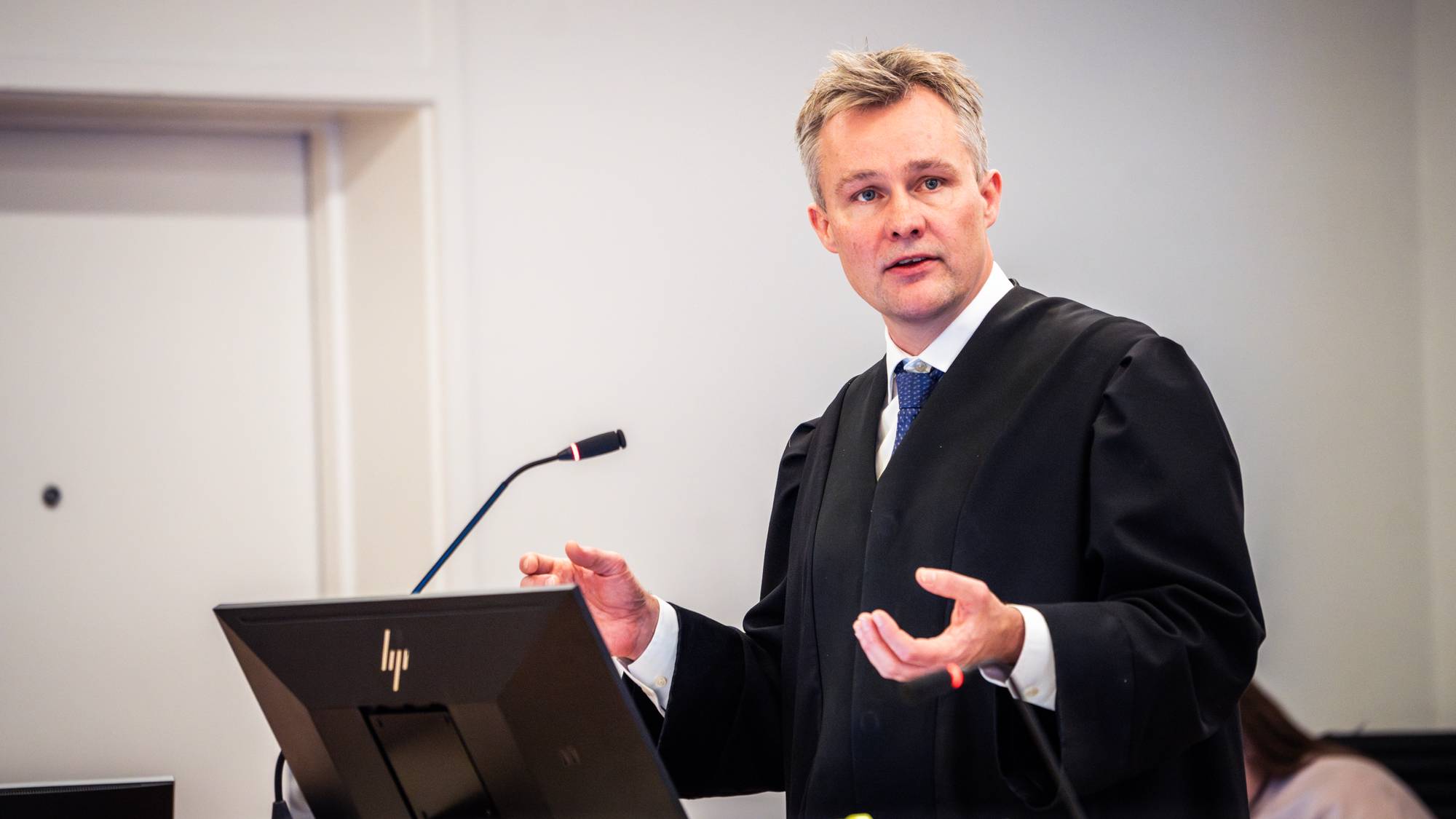 En advokat i svart kappe taler foran en PC-skjerm i et rettslokale. 