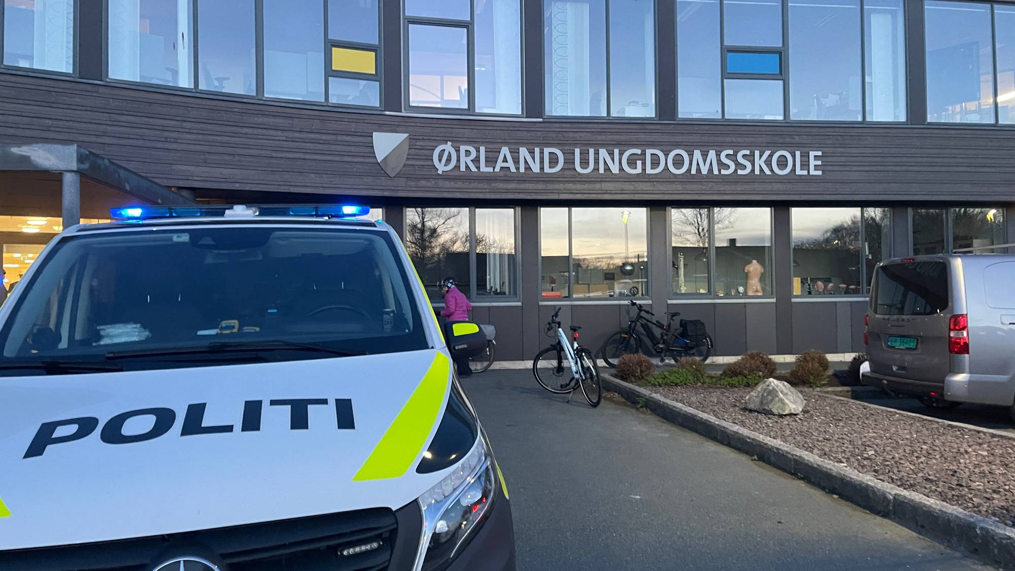 En politibil med blålys står parkert foran Ørland ungdomsskole. Bakgrunnen viser bygget til skolen med store vinduer. To sykler står parkert utenfor skolen. (Bildebeskrivelsen er laget av en KI-tjeneste)