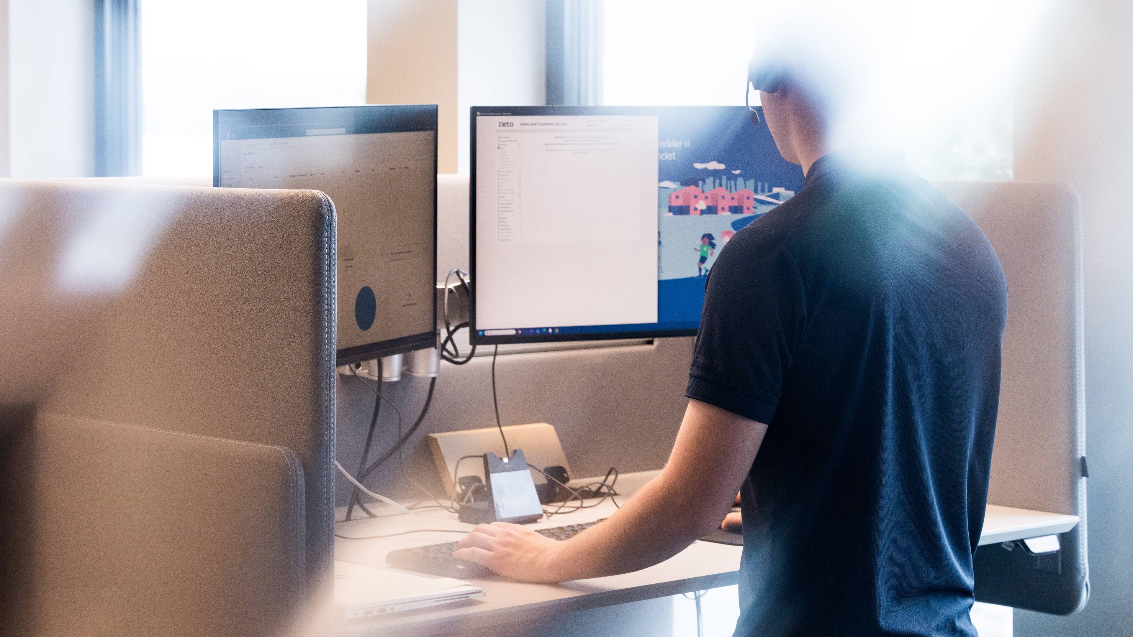 En ung mann står foran en pult med to skjermer. Han har på seg telefonheadset og blå skjorte med korte ermer. Ansiktet er ikke synlig, fordi mannen er anonym. Bildet er mye hvitt lys i seg.