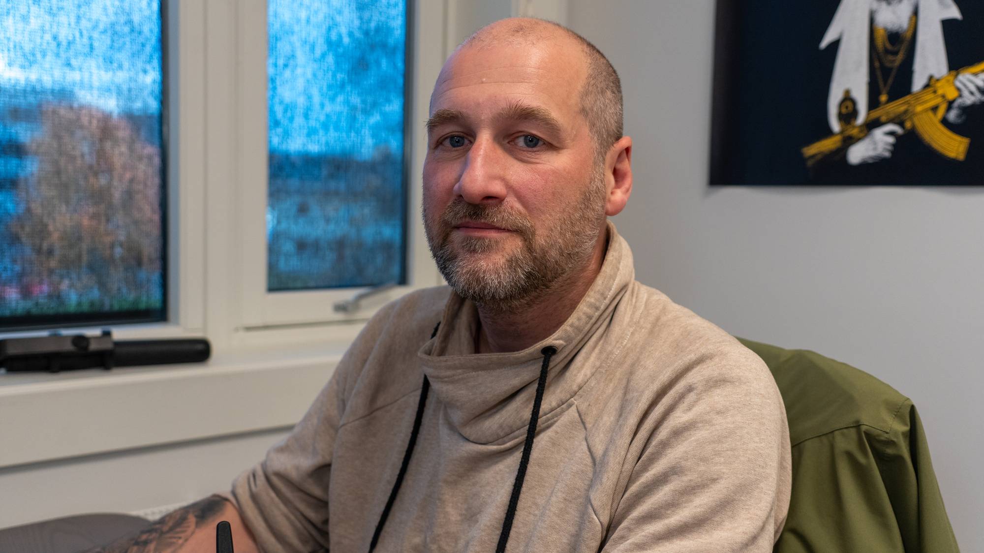En skallet mann med skjegg sitter innendørs ved et vindu. På veggen henger et bilde av en figur. (Bildebeskrivelsen er laget av en KI-tjeneste)