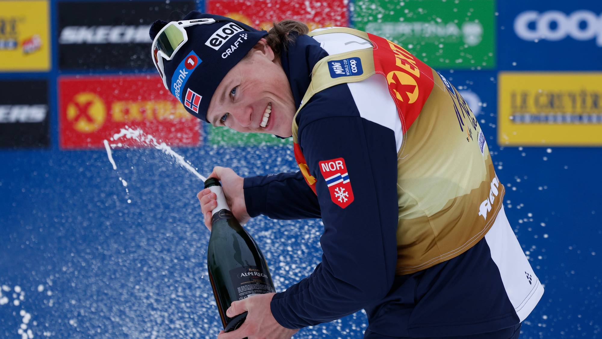 Langrennsløper Johannes Høsflot Klæbo spruter champagne foran en reklamevegg. (Bildebeskrivelsen er laget av en KI-tjeneste)