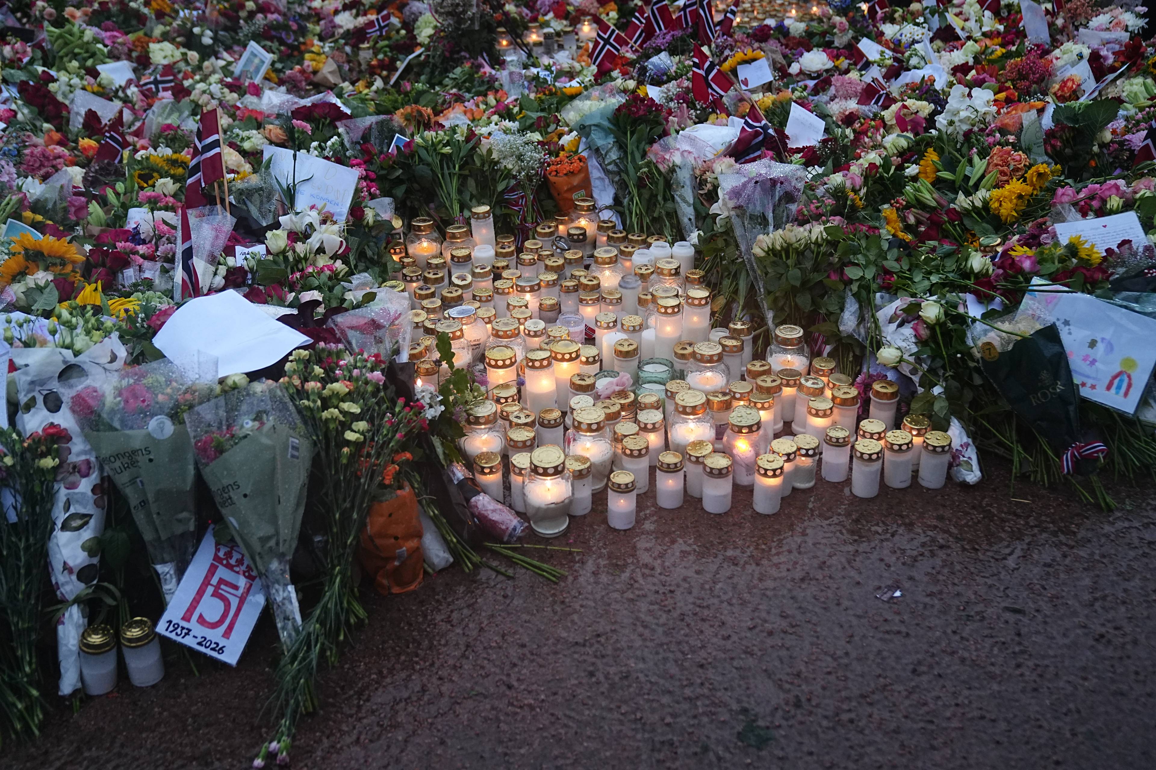 Mange tente lys og blomster lagt ned på et utendørs minnested. Små norske flagg er plassert i havet. (Bildebeskrivelsen er laget av en KI-tjeneste)