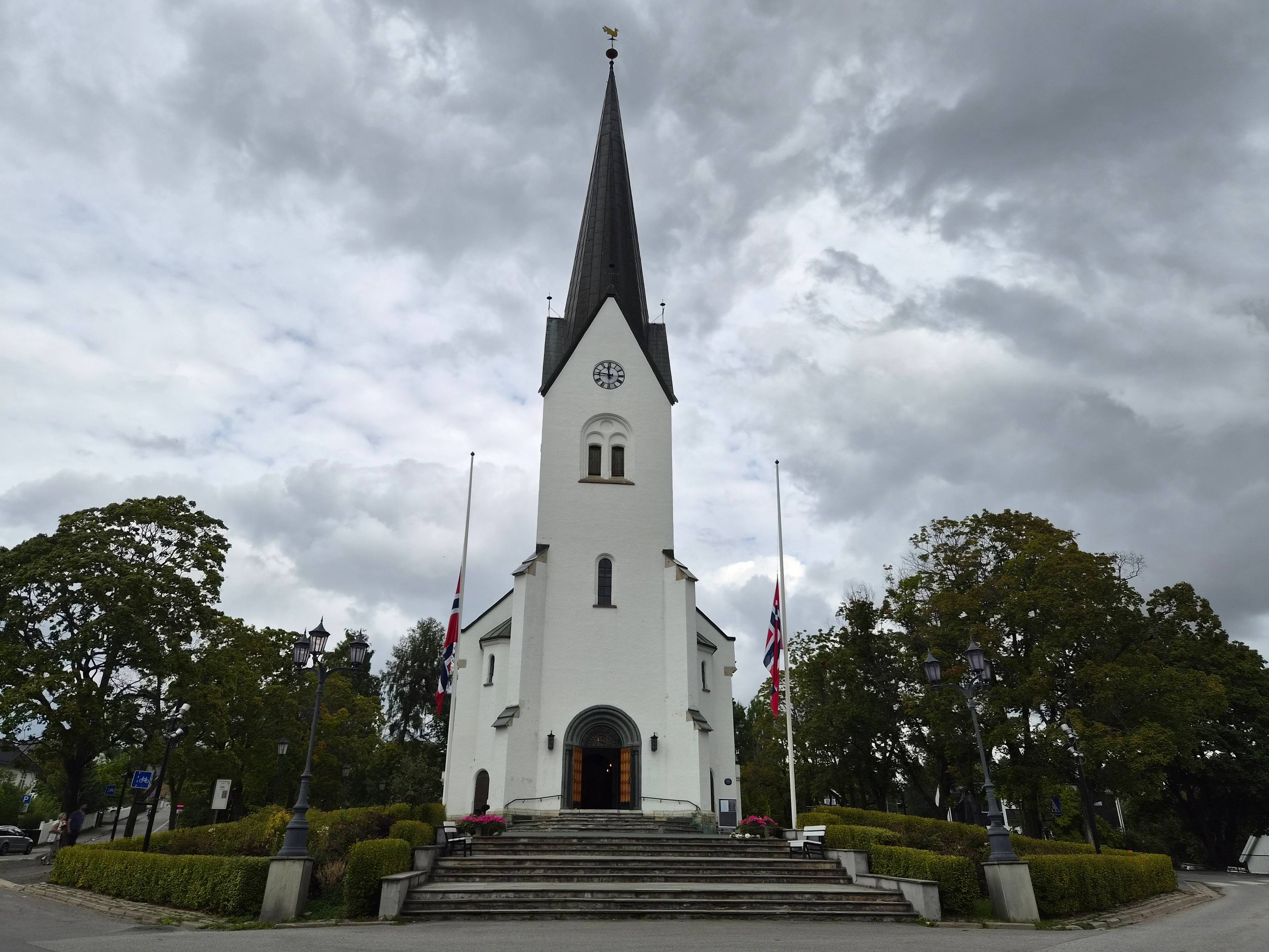 Hvit kirke med to norske flagg på halv stang foran en grå himmel. (Bildebeskrivelsen er laget av en KI-tjeneste)