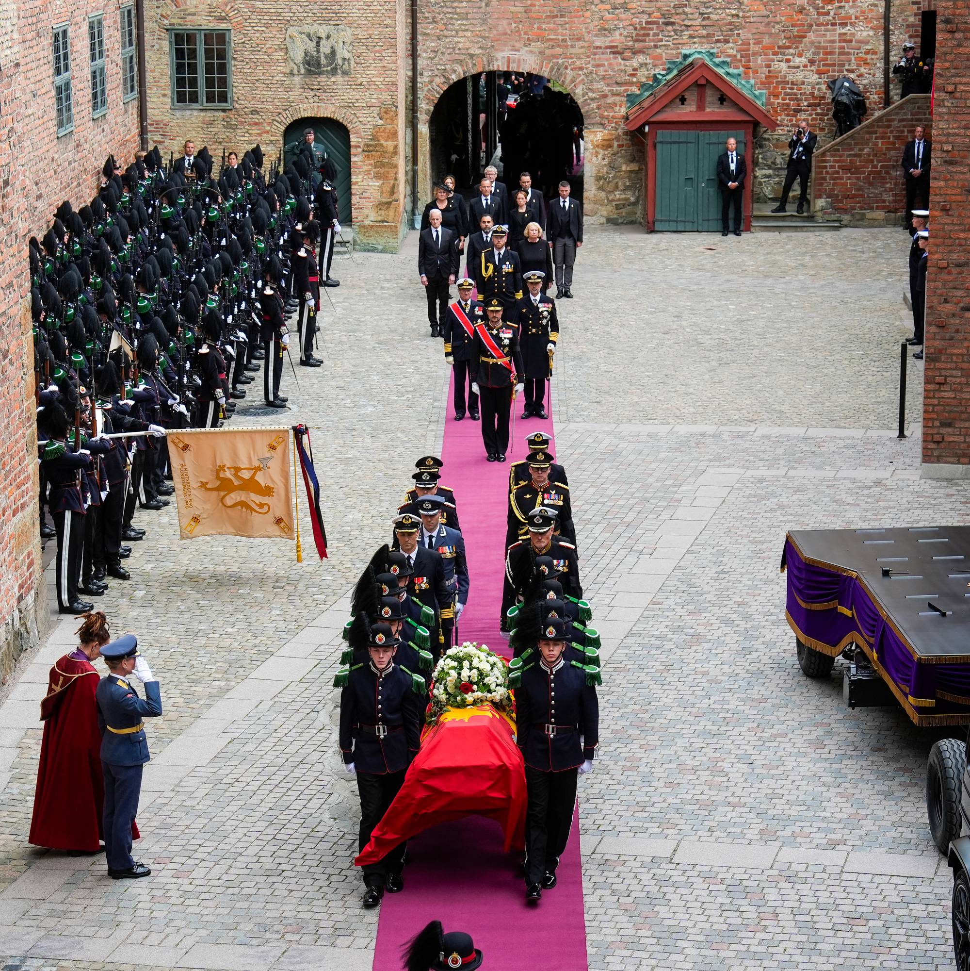 Gardister bærer kong Haralds båre på en fiolett løper inn i Akershus slottskirke. Bak båren går kongens adjutanter, og lenger bak står kong Haakon, kong Carl Gustaf og kong Frederik foran hoffets ledelse.