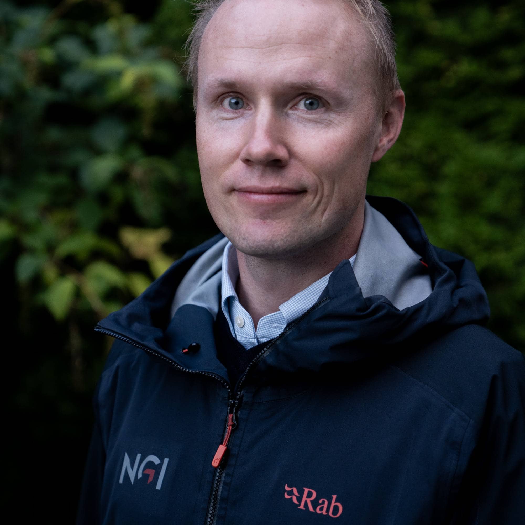 Portrett av en lyshåret mann i mørk jakke med NGI- og Rab-logo, foran en grønn hekk. (Bildebeskrivelsen er laget av en KI-tjeneste)