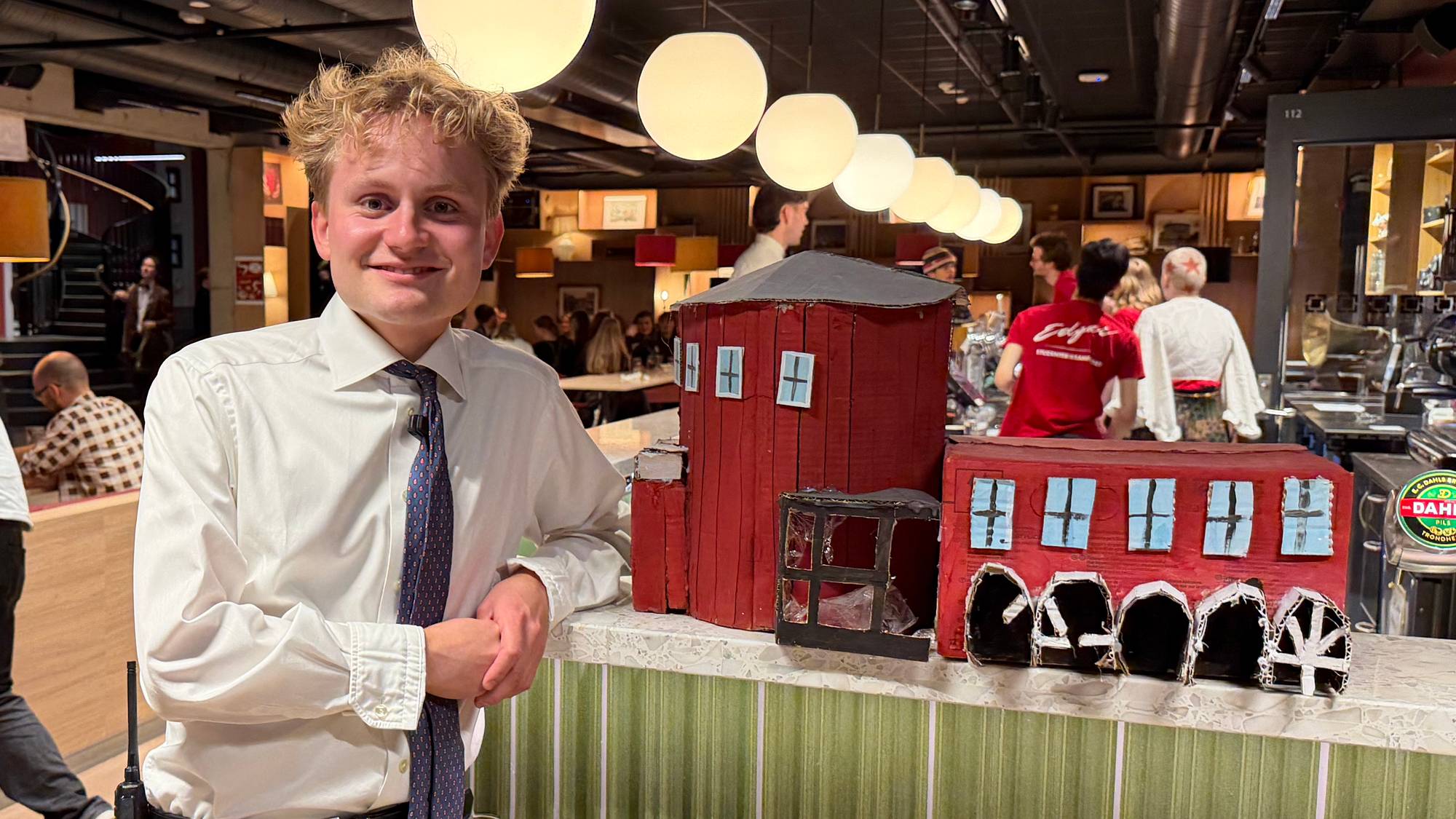 En ung mann i skjorte og slips står ved en bardisk i et lokale med gjester. Ved siden står en modell av Studentersamfundet i papp. (Bildebeskrivelsen er laget av en KI-tjeneste)