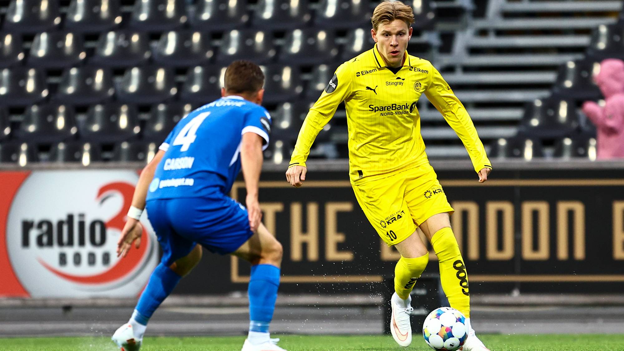 Fotballspiller i gul drakt fører ballen foran en motstander i blå drakt på en fotballbane. (Bildebeskrivelsen er laget av en KI-tjeneste)