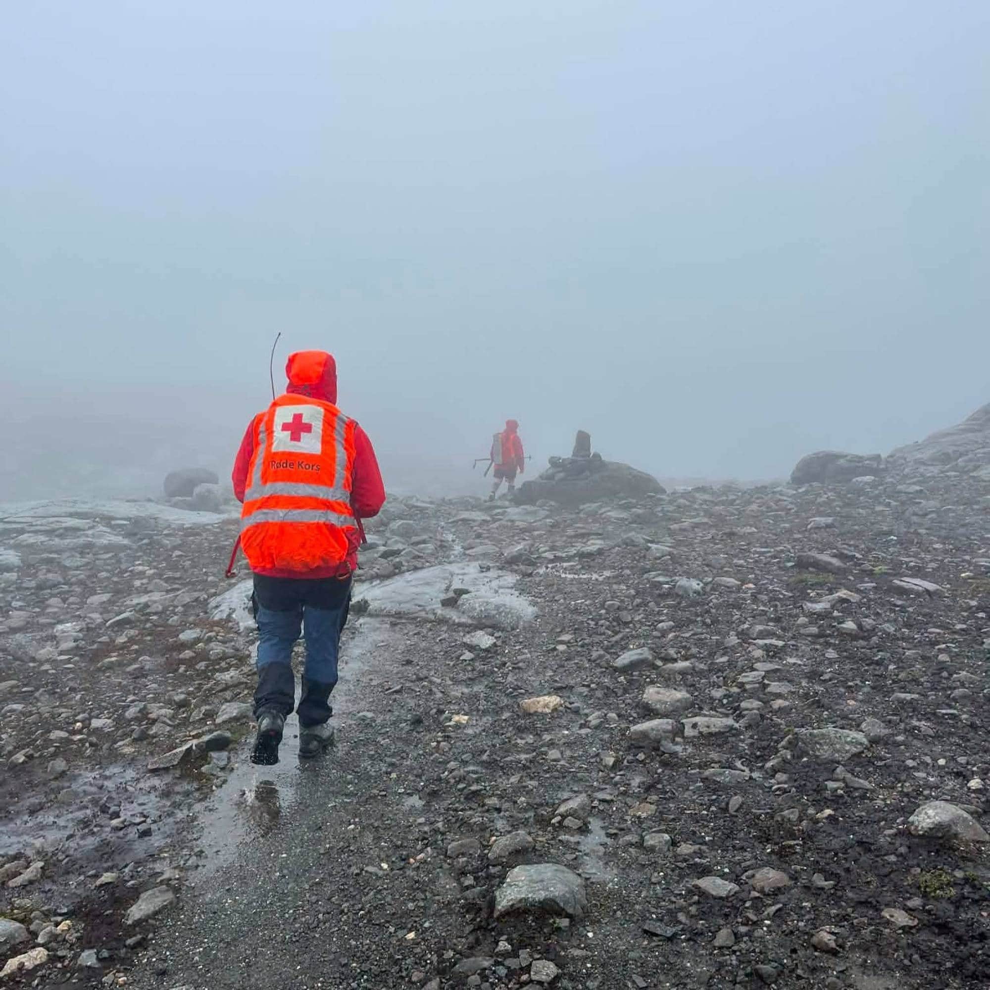 Person i oransje kle og røde kors merke går i tåke på fjellet.