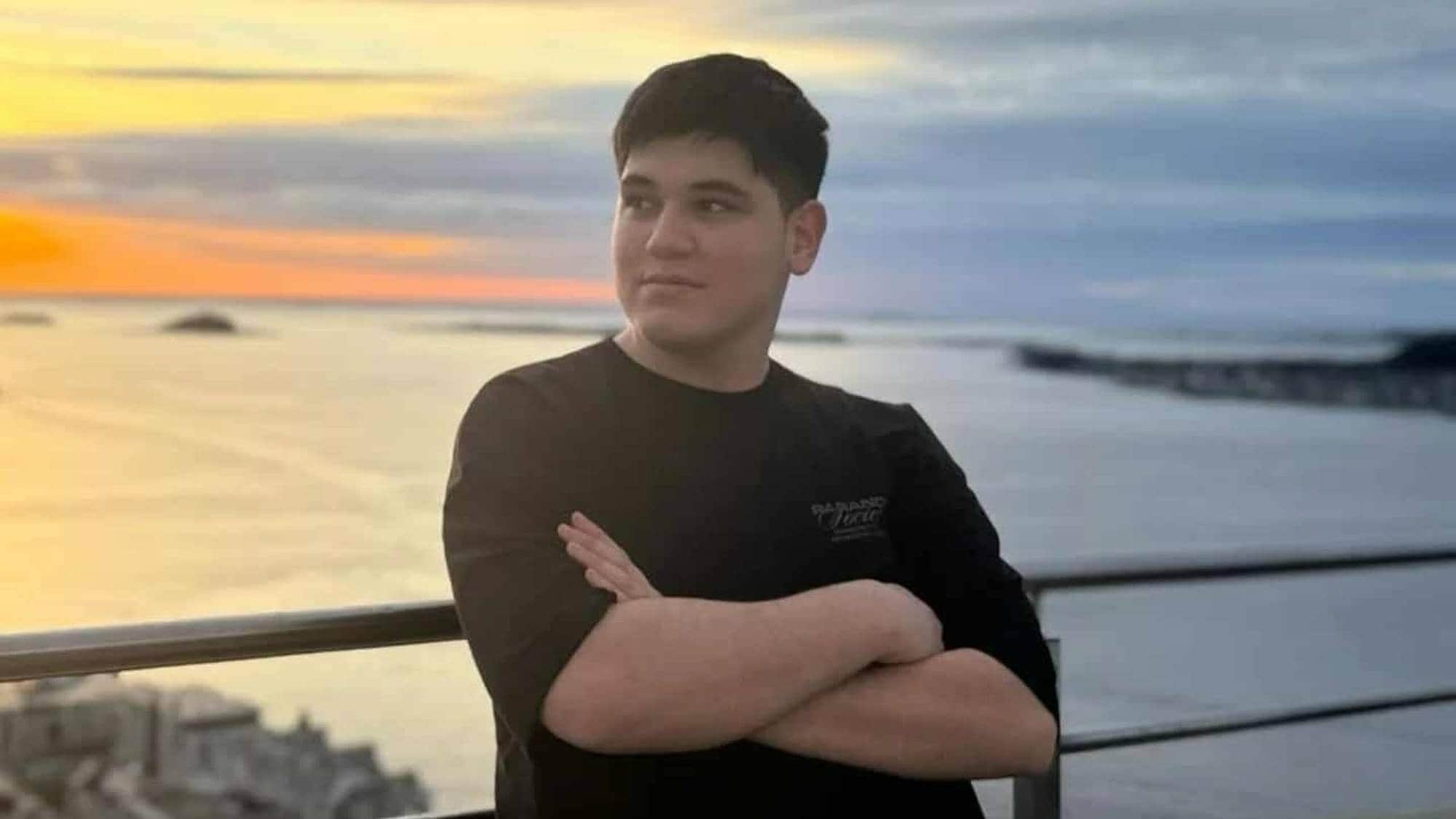 En person står med armene krysset foran seg, mens de ser utover havet. Solnedgangen farger himmelen i gule og oransje nyanser. Bakgrunnen viser en kystlinje og bygninger. Bildet gir inntrykk av et rolig øyeblikk ved sjøen.