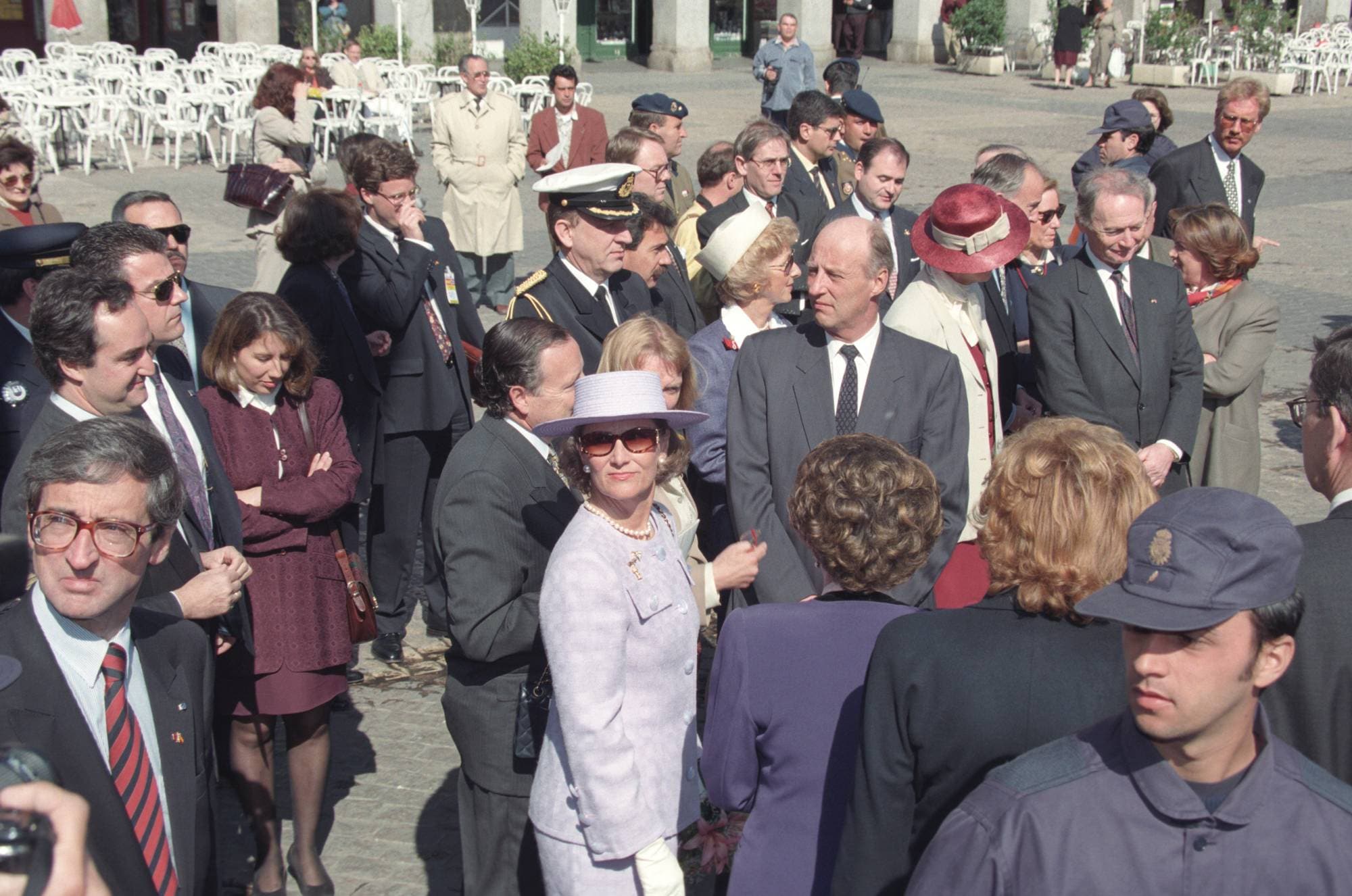 Dronning Sonja i lys drakt og hatt omgitt av en folkemengde i dress og formelle klær på en plass. (Bildebeskrivelsen er laget av en KI-tjeneste)