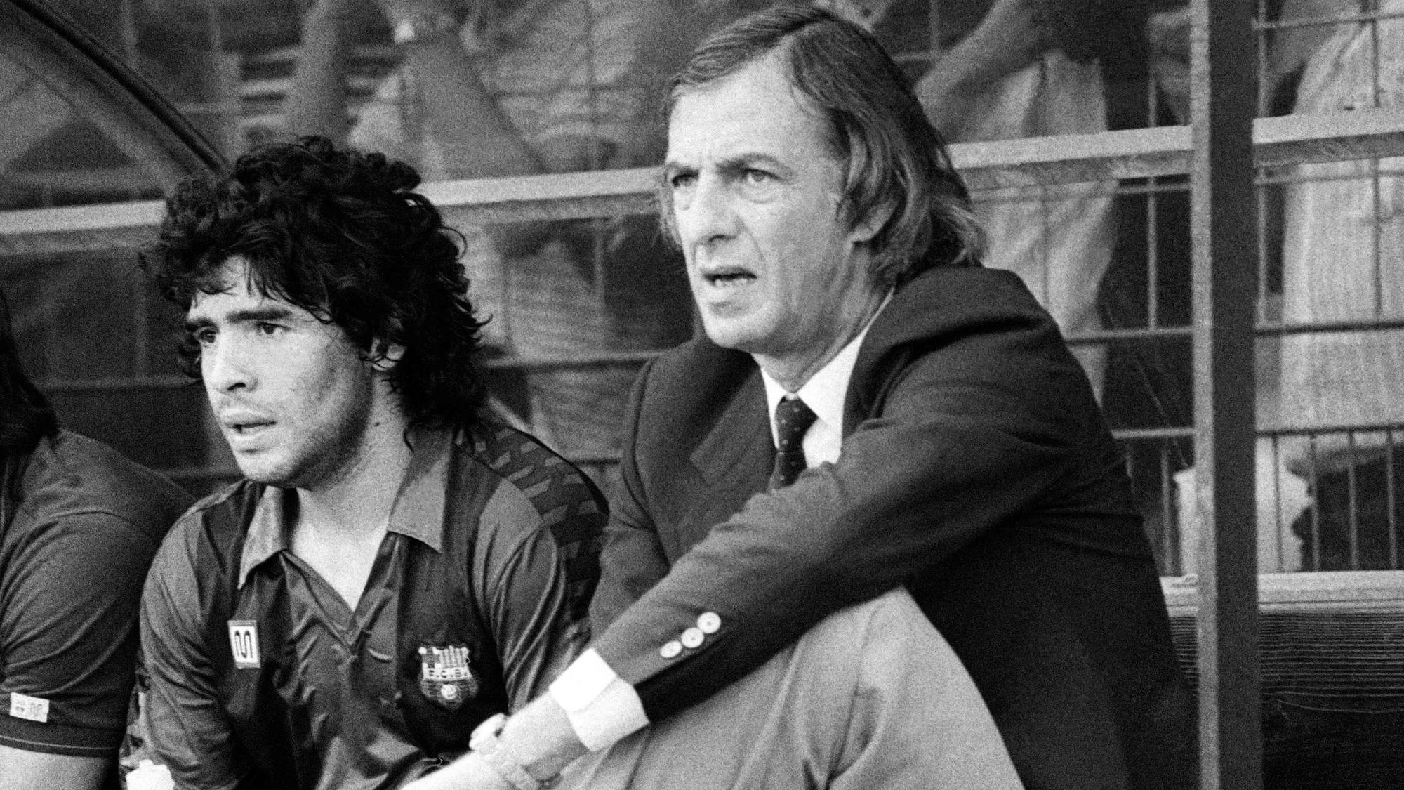 Tre menn sitter på en benk. Mannen i midten, Diego Maradona, ser ut av bildet. Mannen til høyre har på seg dressjakke og slips. Mennene sitter på en sportsarena, med tilskuere i bakgrunnen. (Bildebeskrivelsen er laget av en KI-tjeneste)