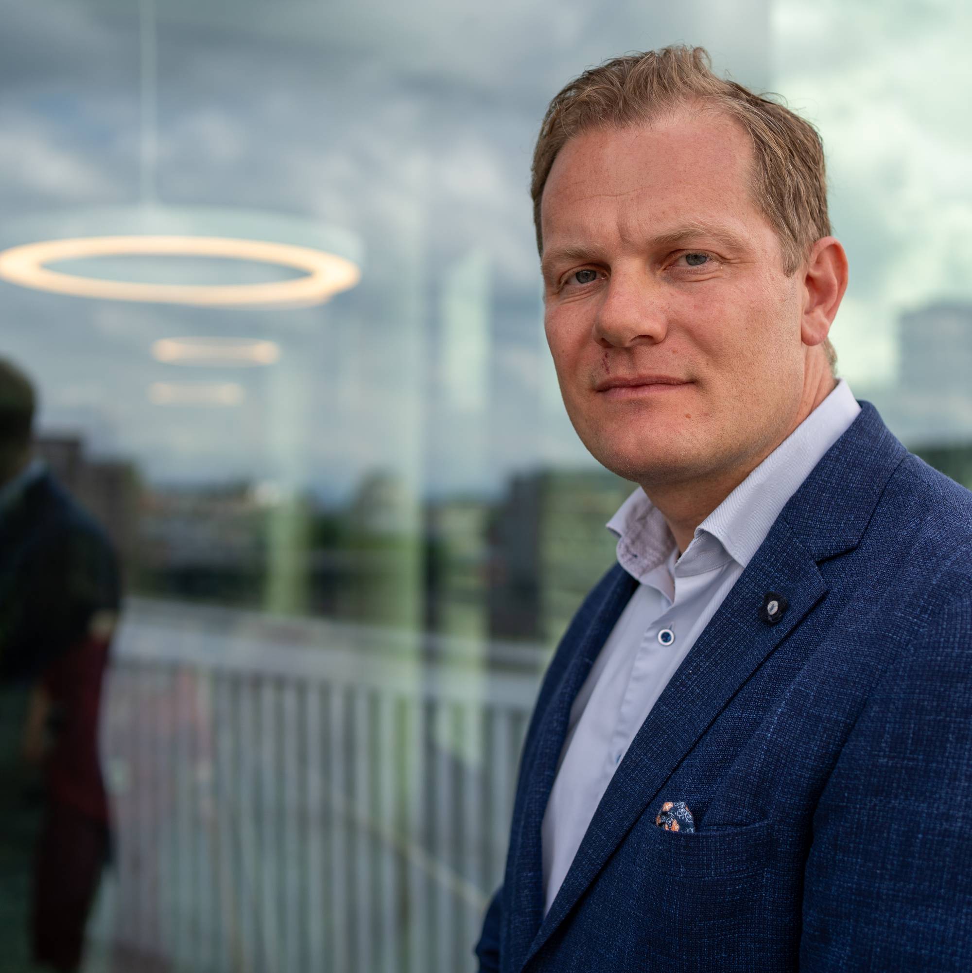 Middelaldrende mann i blå dress og lyseblå skjorte står til høyre i bildet foran en glassvegg. Glassveggen viser inn til et kantineområde bak, men viser også gjenskinn av himmelen. Mannen ser i kamera.