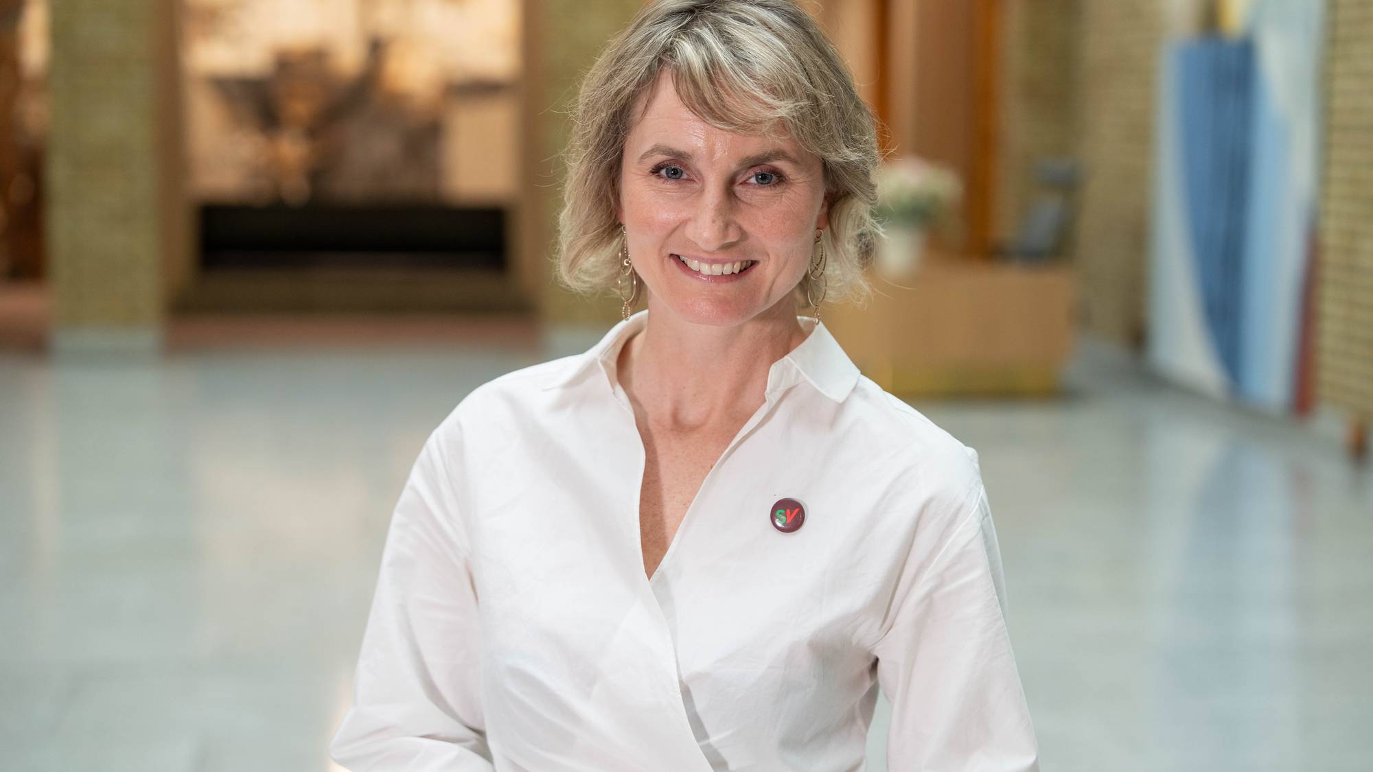 En kvinne med blondt hår og et smil poserer foran et kamera. Hun har på seg en hvit skjorte med en liten rund knapp med grønn og rød tekst på venstre side av brystet. Bakgrunnen er uskarp, men viser en bygning med lyse vegger og noe som kan være kunst på veggen. (Bildebeskrivelsen er laget av en KI-tjeneste)