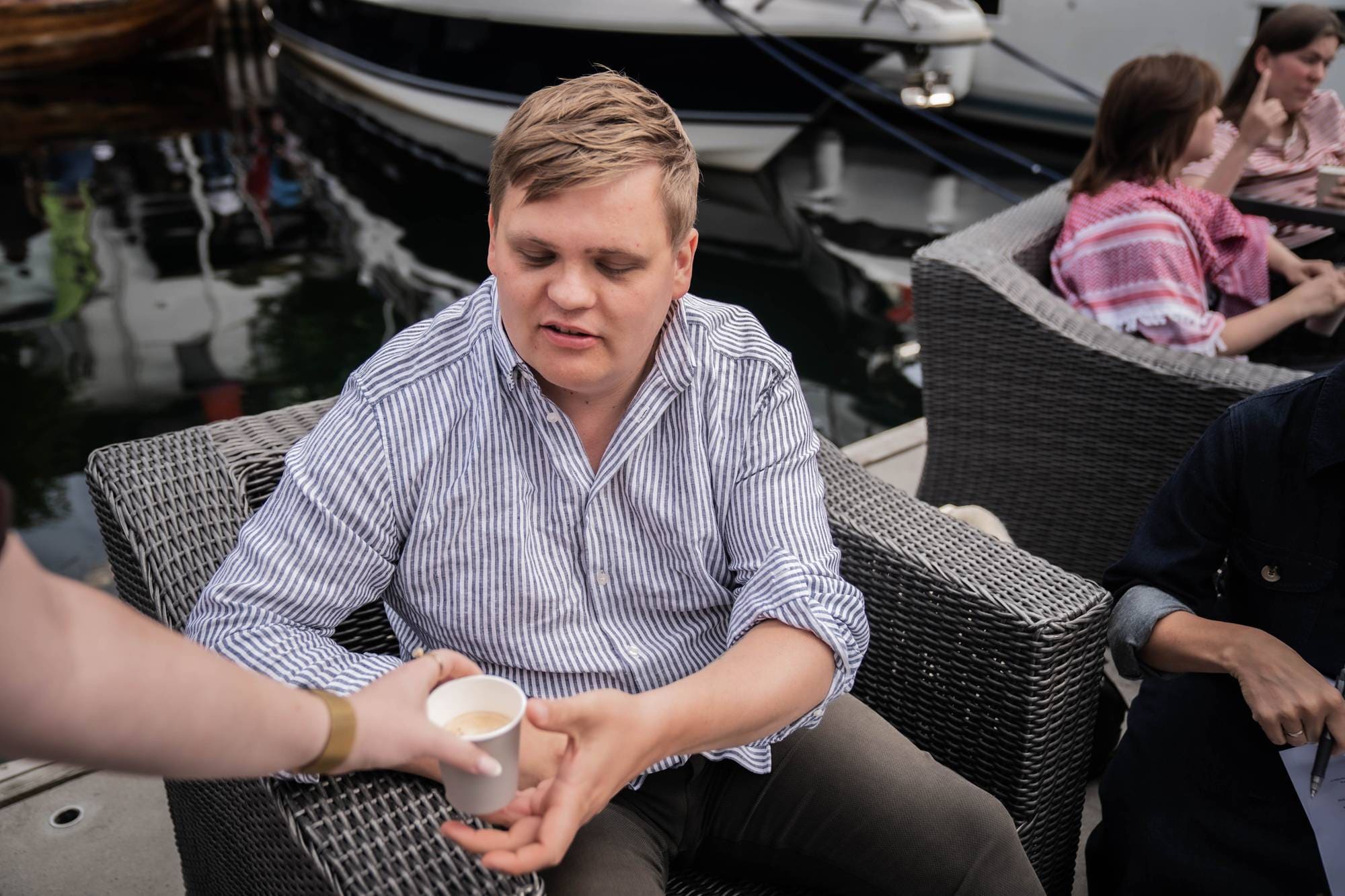Mann i stripete skjorte sitter i en stol ved en båthavn og mottar en kaffekopp. (Bildebeskrivelsen er laget av en KI-tjeneste)