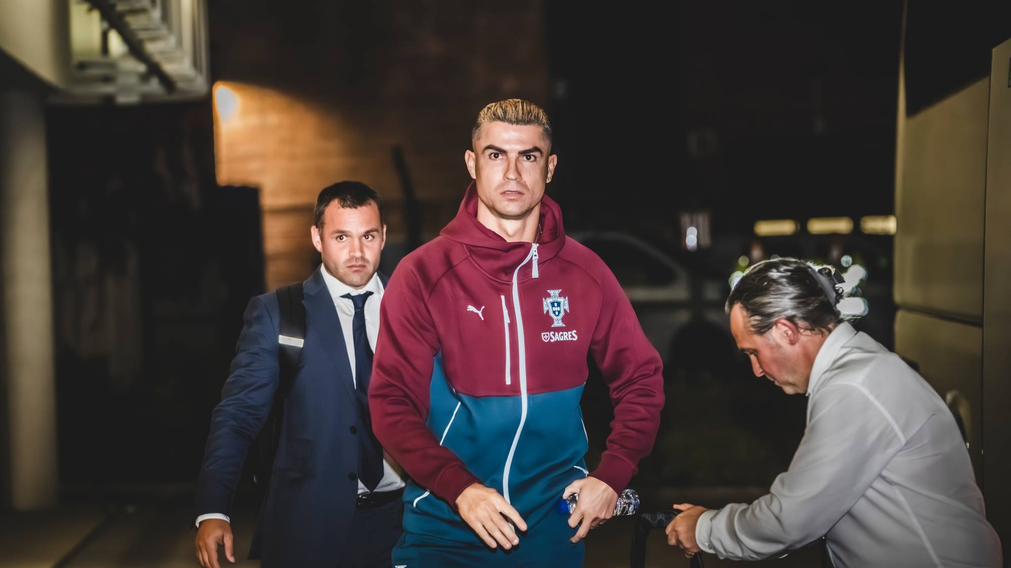 Cristiano Ronaldo går utendørs kledd i Portugals landslagstøy, flankert av to menn i dress.