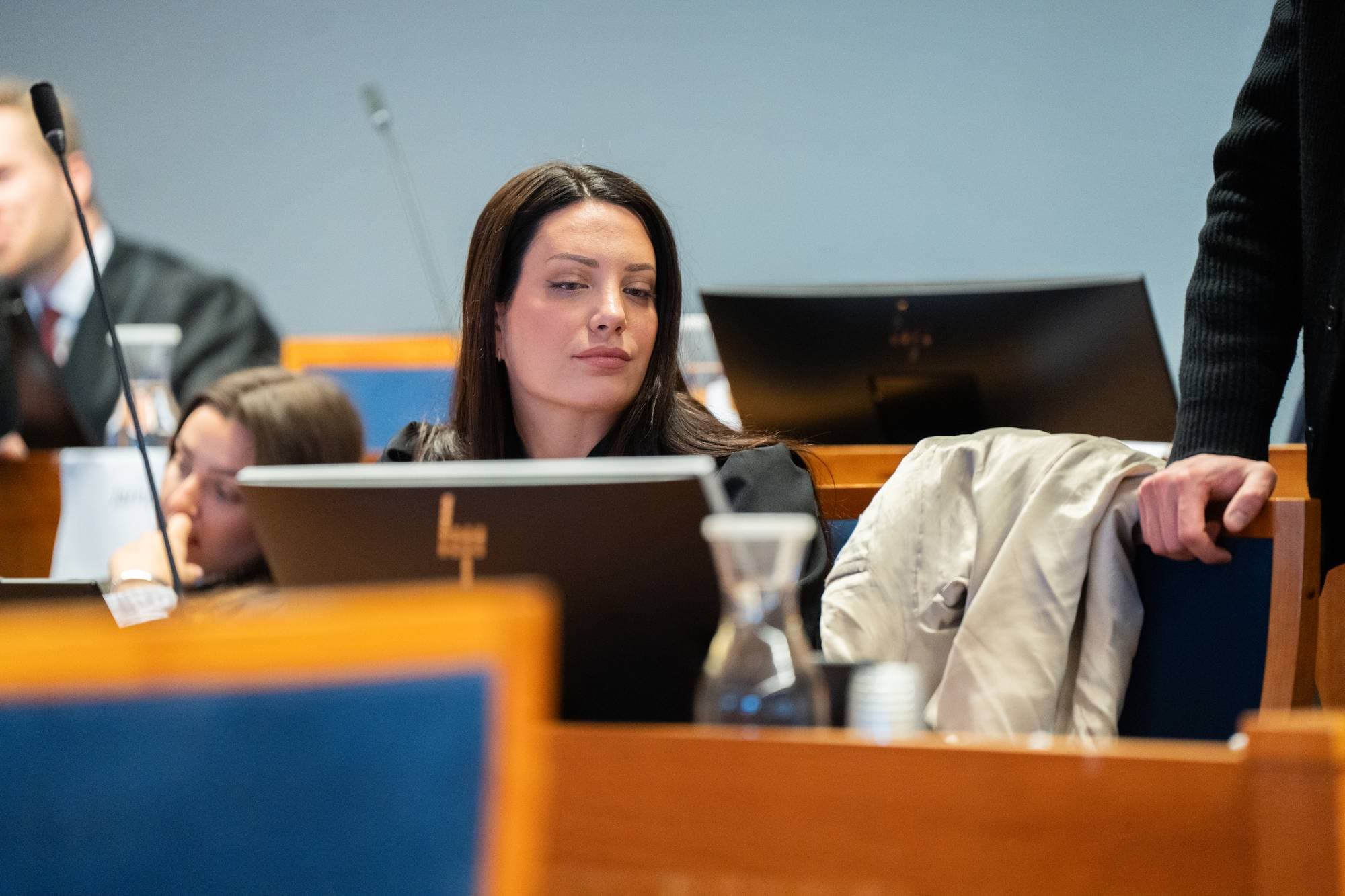 En kvinne sitter i rettssal, foran en skjerm. Hun er flankert av andre personer.
Bildet viser en scene fra en rettssal. Kvinnen ser mot høyre, mens en mann står lent mot stolen ved siden av henne.
En annen person, en kvinne, sitter til venstre for hovedpersonen i bildet.
