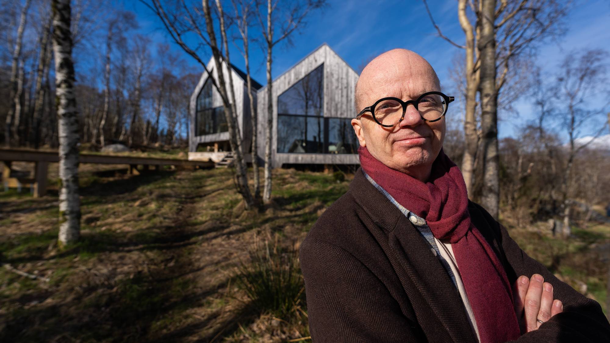 Mann med briller og skjerf utenfor et moderne hus. Huset er en arkitekttegnet trekonstruksjon med store vindusflater. Mannen ser mot kamera og har armene i kors. Bakgrunnen består av trær og en blå himmel. (Bildebeskrivelsen er laget av en KI-tjeneste)