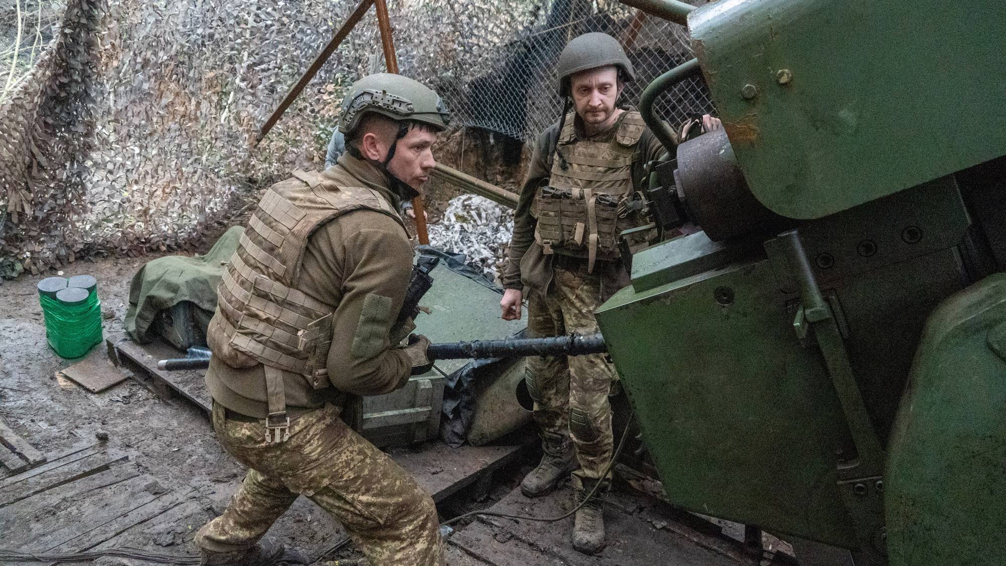 To soldater i kamuflasjeklær og med hjelm arbeider ved et grønt artilleristykke. En soldat holder i en jernstang som er festet til kanonen. Den andre soldaten ser på.Bak dem er det kamuflasjenett, som gir kontekst til en militær stilling. (Bildebeskrivelsen er laget av en KI-tjeneste)