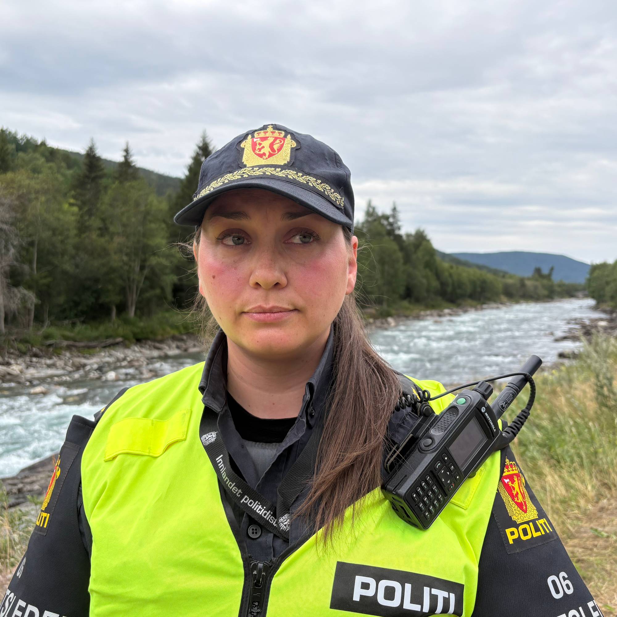 En politibetjent i uniform står utendørs med en elv og skog i bakgrunnen. 