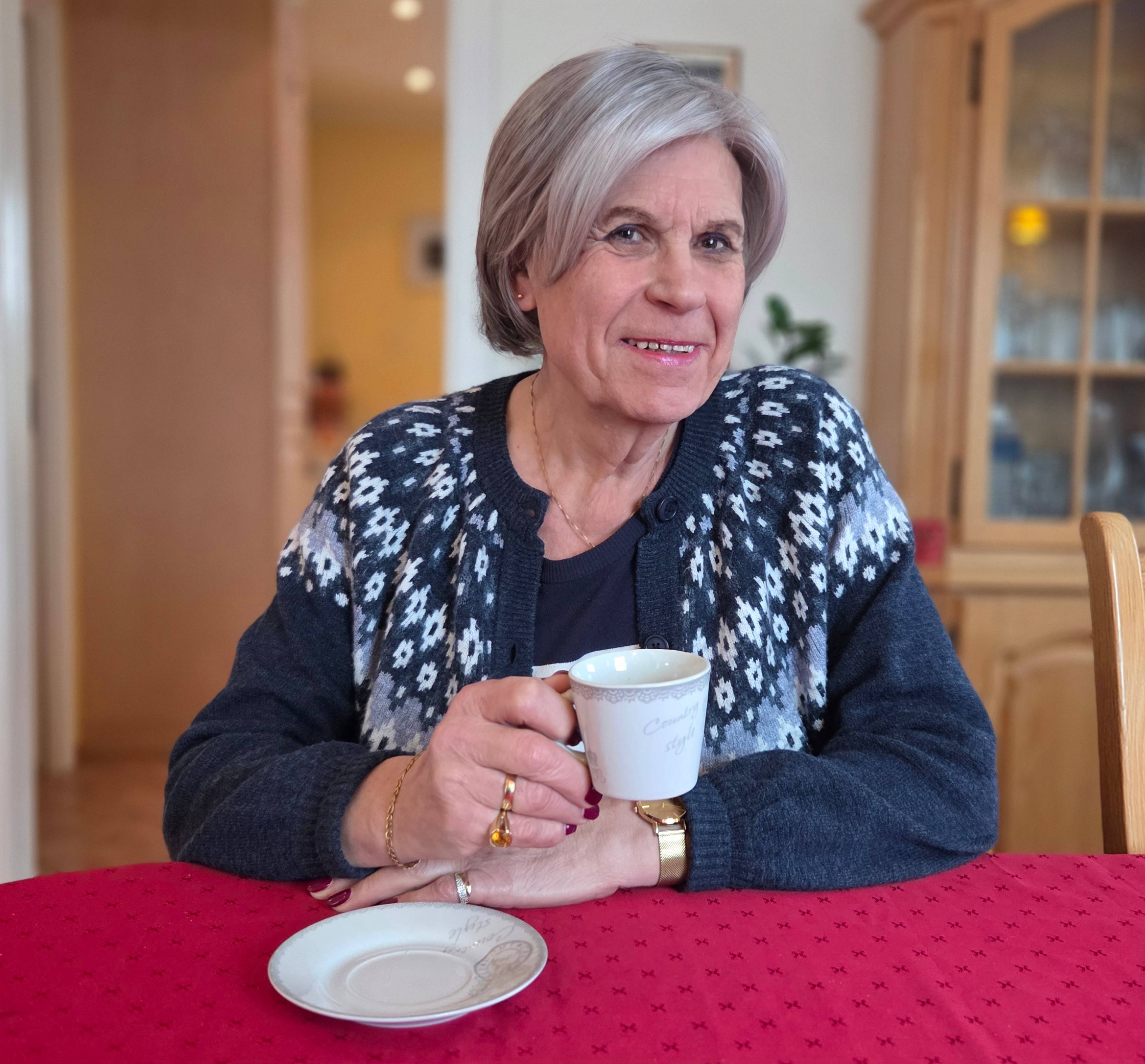 En eldre transperson sitter og smiler ved et bord med rød duk og drikker kaffe. Hun har grå parykk og strikkegenser.
