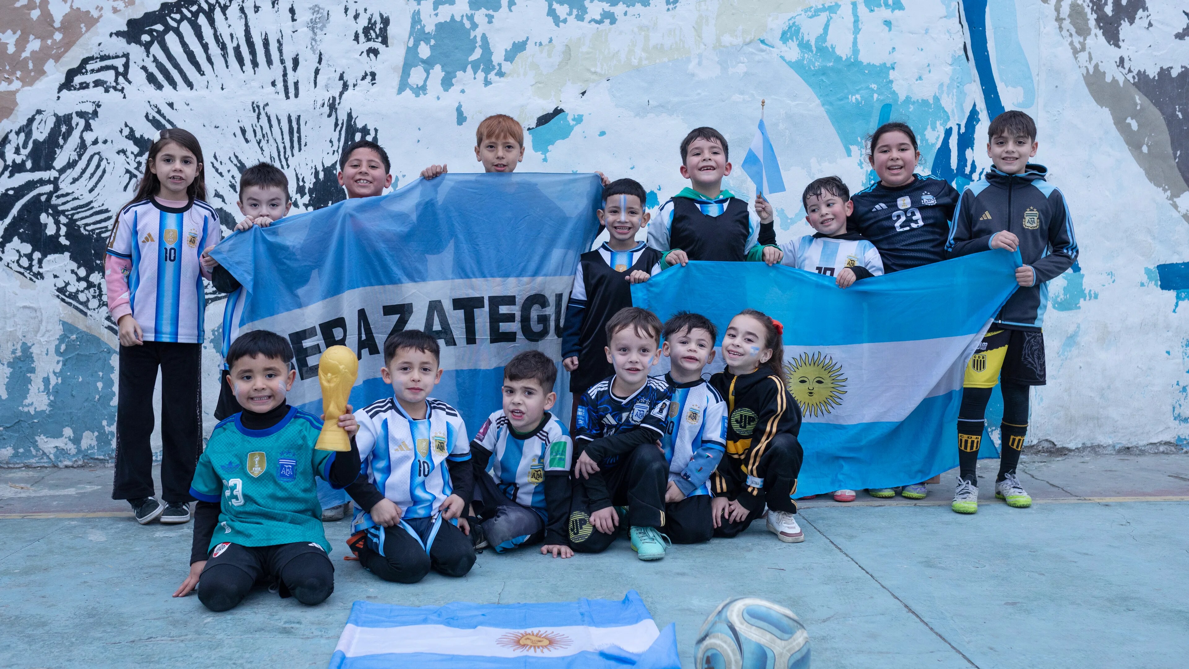 En gruppe barn i Argentina-drakter holder flagg og en VM-pokal foran en malt vegg.