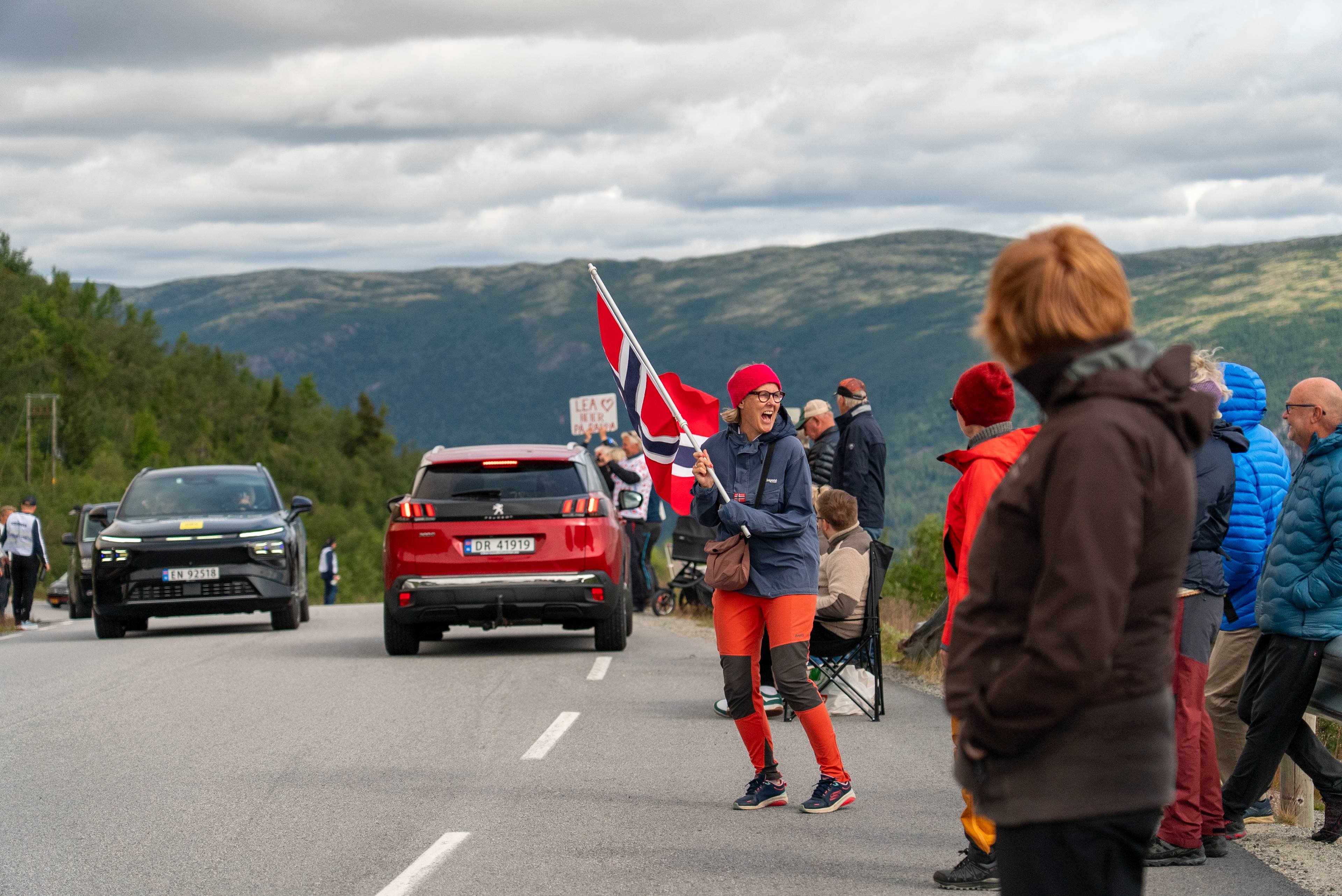 Kvinne med norsk flagg heier langs en vei med biler i et fjellandskap. (Bildebeskrivelsen er laget av en KI-tjeneste)