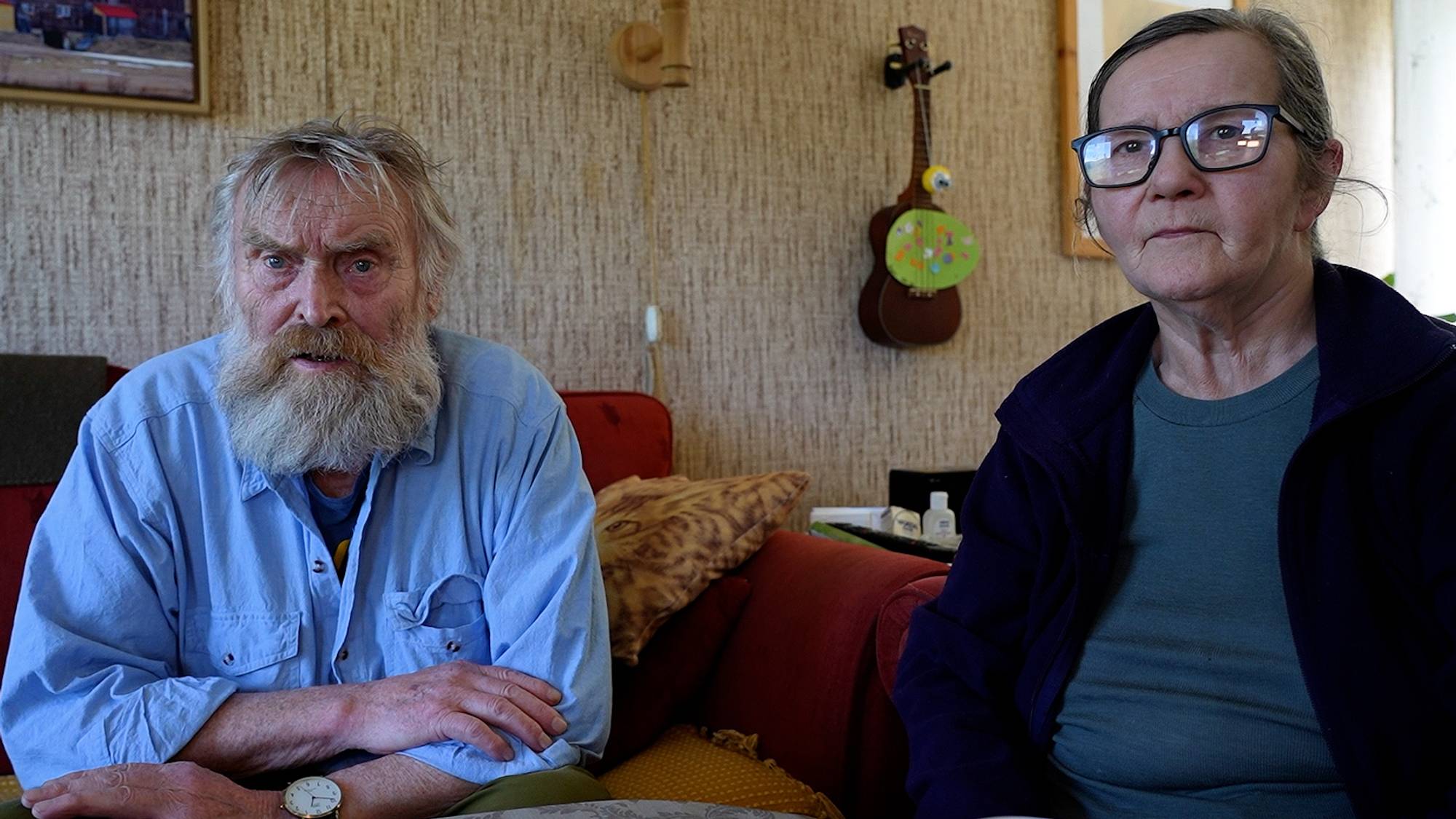 En eldre mann med skjegg og en eldre kvinne med briller sitter sammen i en stue. (Bildebeskrivelsen er laget av en KI-tjeneste)