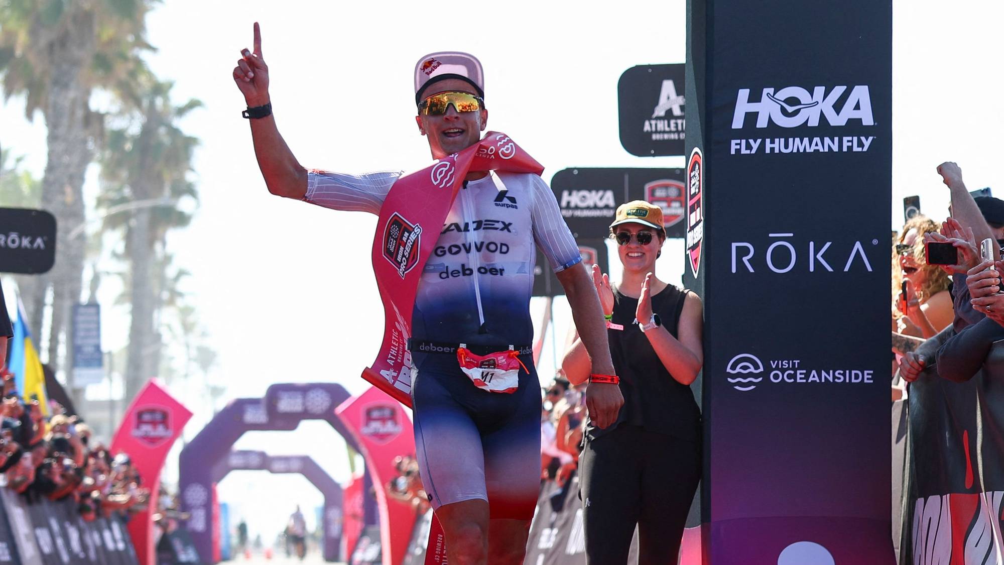 En mann i en triatlon-drakt krysser mållinjen. Han peker opp med høyre hånd og har en rød tøyduk over skuldrene. En kvinne med solbriller og caps klapper til høyre for mannen. Til venstre står en mann i en mørk t-skjorte og mørke shorts og klapper. Bak dem er det publikum, tribuner og bueganger. (Bildebeskrivelsen er laget av en KI-tjeneste)