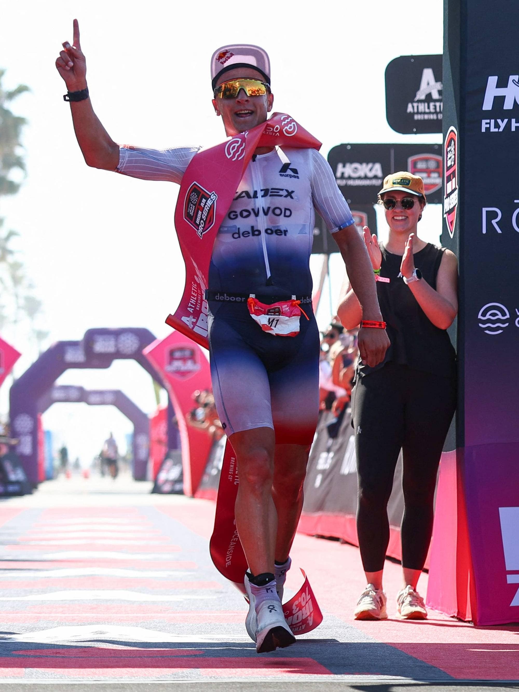 En mann i en triatlon-drakt krysser mållinjen. Han peker opp med høyre hånd og har en rød tøyduk over skuldrene. En kvinne med solbriller og caps klapper til høyre for mannen. Til venstre står en mann i en mørk t-skjorte og mørke shorts og klapper. Bak dem er det publikum, tribuner og bueganger. (Bildebeskrivelsen er laget av en KI-tjeneste)