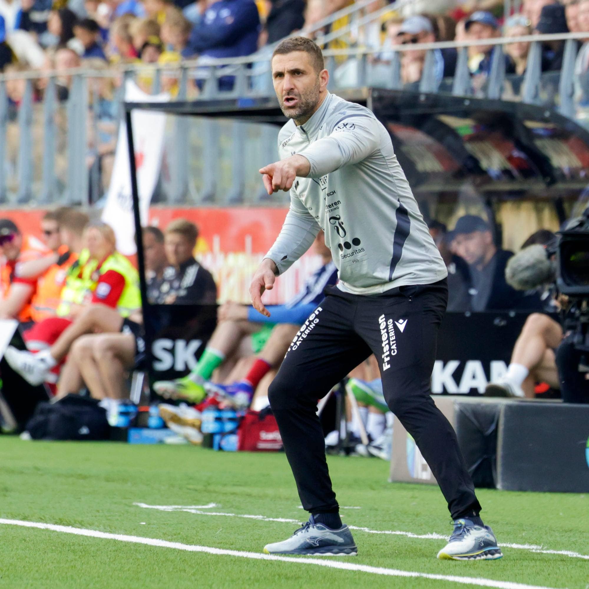 En fotballtrener peker og snakker. Han står på en gressmatte. Bak ham sitter folk på tribunen og i et busskur. En TV-kameramann observerer. (Bildebeskrivelsen er laget av en KI-tjeneste)