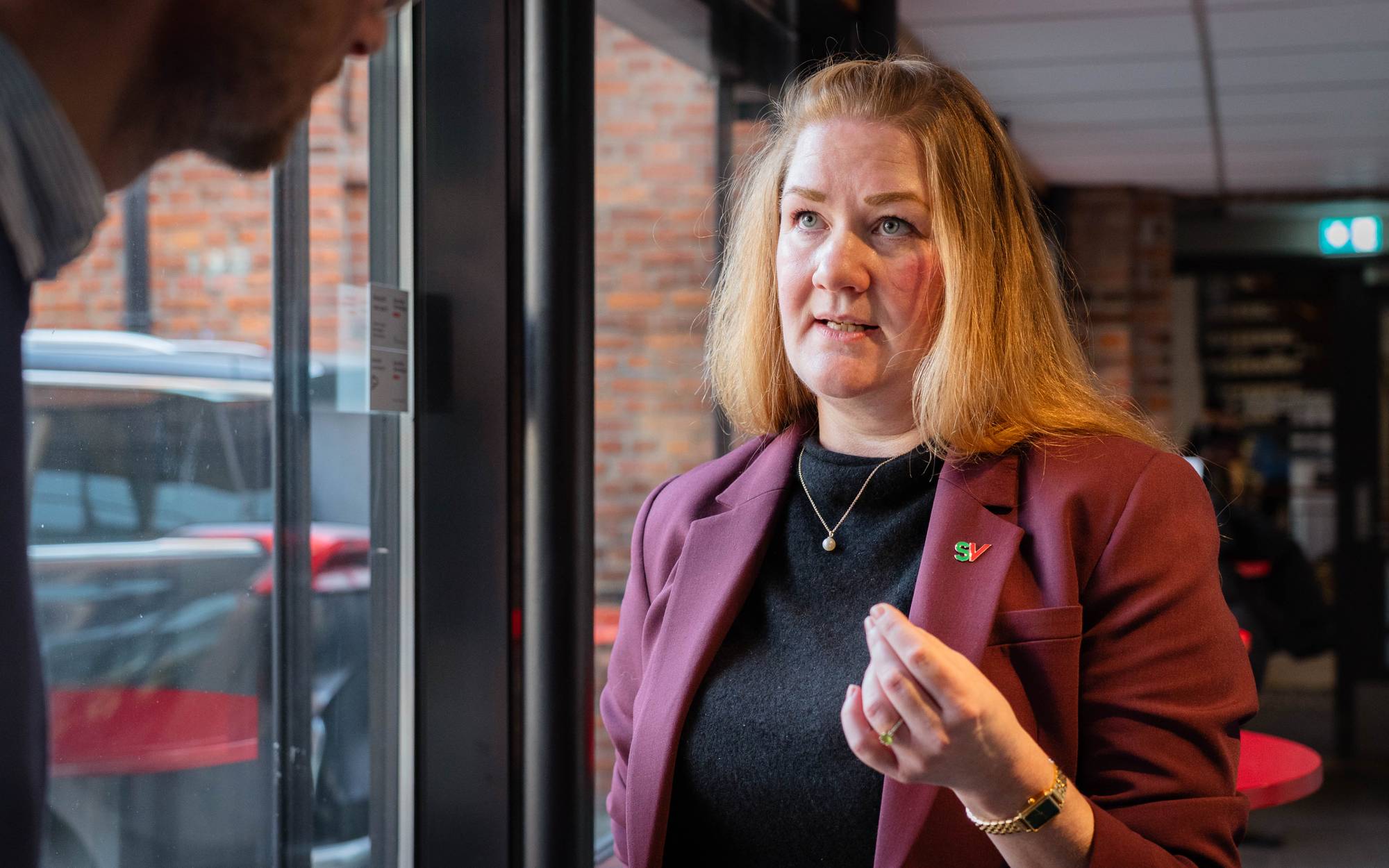 Bildet viser en person i en rød blazer som står og gestikulerer, tydelig engasjert i en samtale. Bakgrunnen inneholder murvegger og møbler som antyder en kafé- eller kontorinnstilling. Det er flere personer og biler synlige i bakgrunnen, men de er ikke i fokus. Det er naturlig lys som kommer inn gjennom vinduene. (Bildebeskrivelsen er laget av en KI-tjeneste)