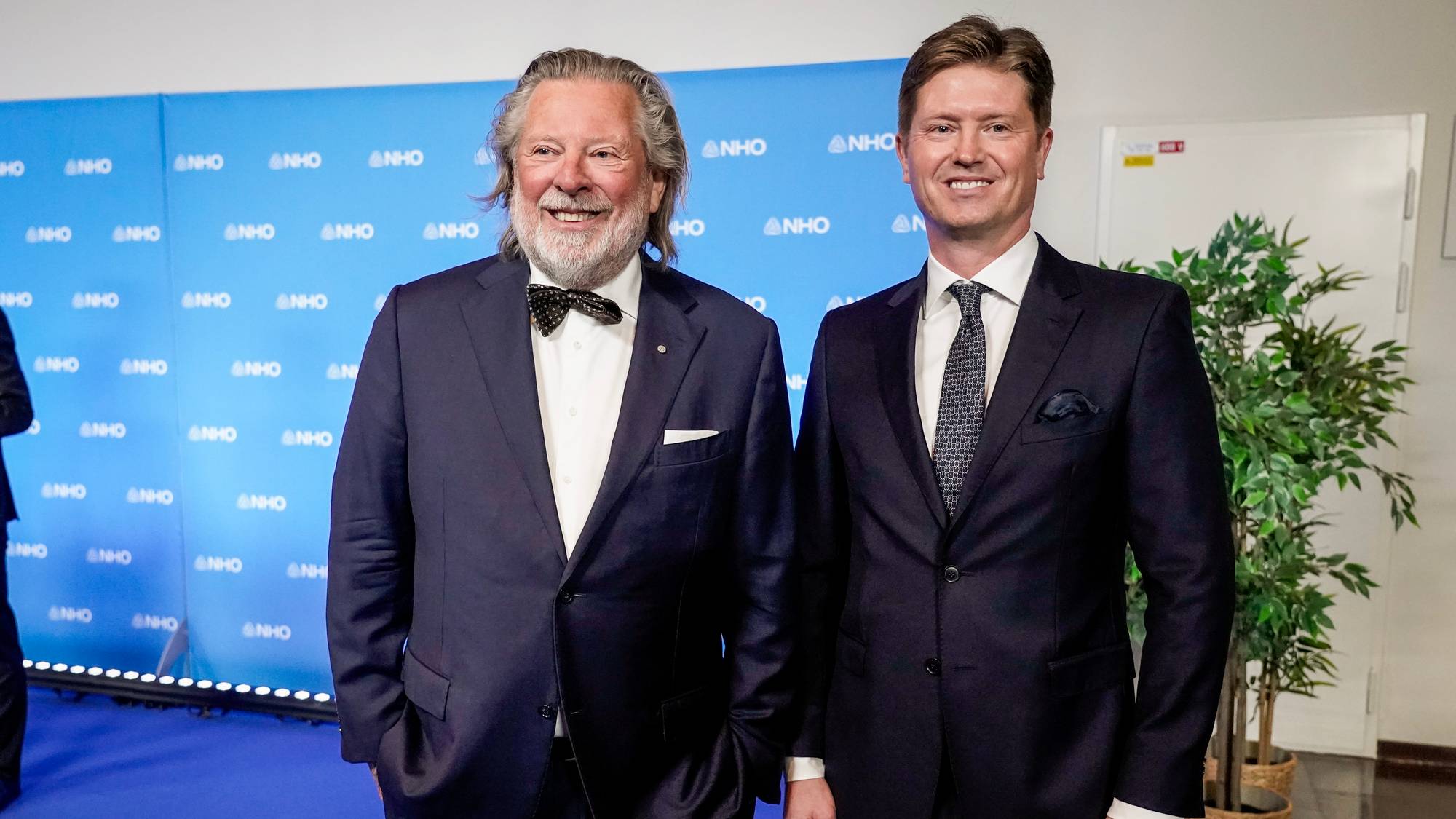 To menn i dress står foran en blå bakgrunn med NHO-logoer. Den ene mannen, som er eldre, smiler bredt. Den andre, yngre mannen, smiler også. Til høyre for dem står en stor grønn plante i en kurvpotte. Begge mennene er kledd i mørke dresser og ser mot kamera. (Bildebeskrivelsen er laget av en KI-tjeneste)