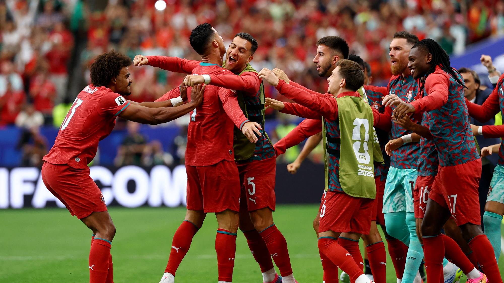 En gruppe fotballspillere i røde drakter feirer sammen på en gressbane. Spillere omfavner hverandre mens de ser opp og smiler. I bakgrunnen kan man se tribuner fylt med tilskuere. (Bildebeskrivelsen er laget av en KI-tjeneste)