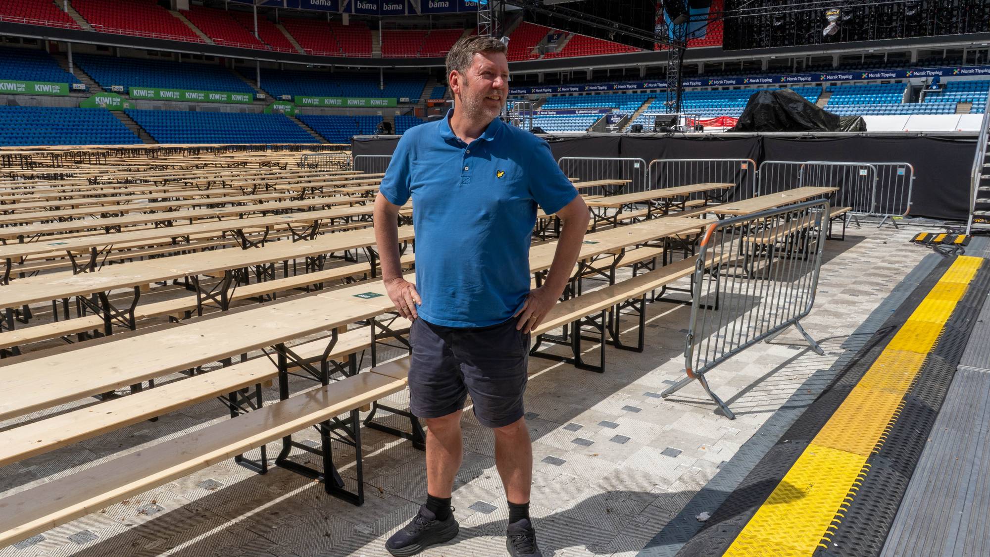 Mann i blå skjorte står på et tomt stadion med mange rader av trebenker foran en scene.