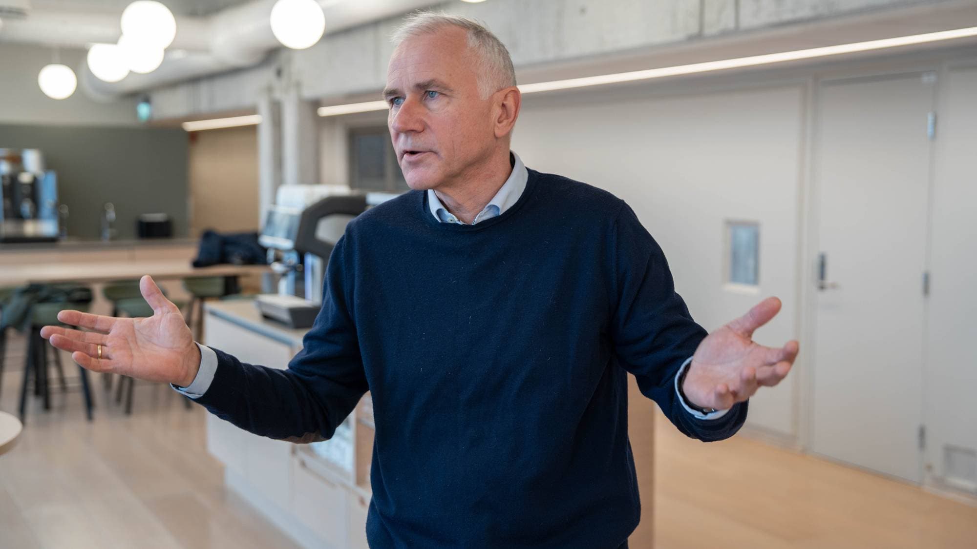 En mann iført en mørk genser gestikulerer med hendene i et moderne rom. Bakgrunnen viser en kaffe- og loungeområde med møbler. Det er godt lys og en moderne interiørdesign. Mannens kroppsspråk indikerer at han tar del i en samtale eller presentasjon.