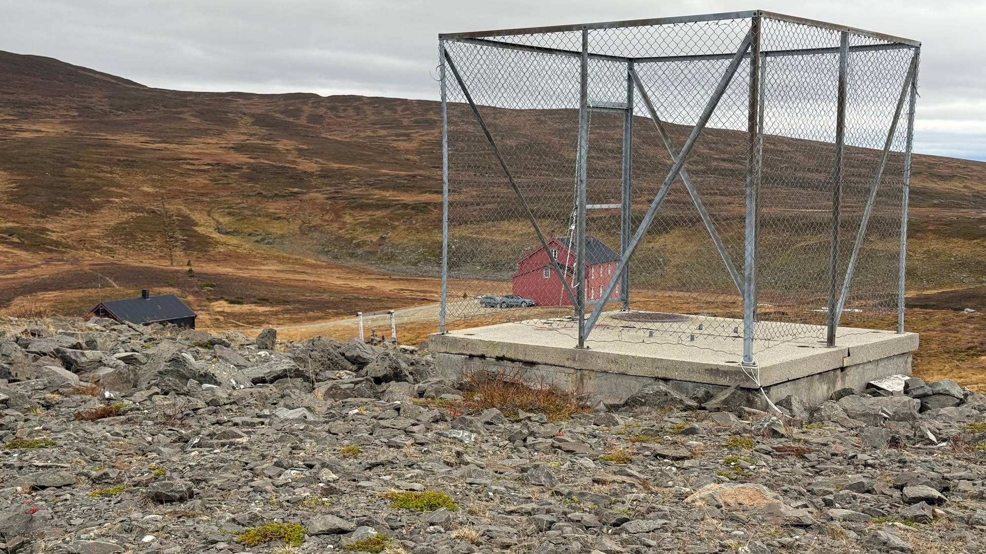 Inngjerdet betongfundament i et fjellandskap med to røde trebygninger i bakgrunnen. Gjerdene er høye. Innenfor gjerdene er et kumlokk. (Bildebeskrivelsen er laget av en KI-tjeneste)