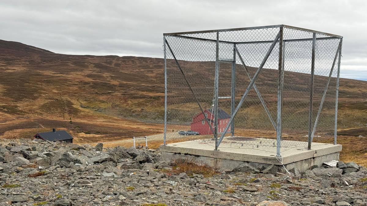 Inngjerdet betongfundament i et fjellandskap med to røde trebygninger i bakgrunnen. Gjerdene er høye. Innenfor gjerdene er