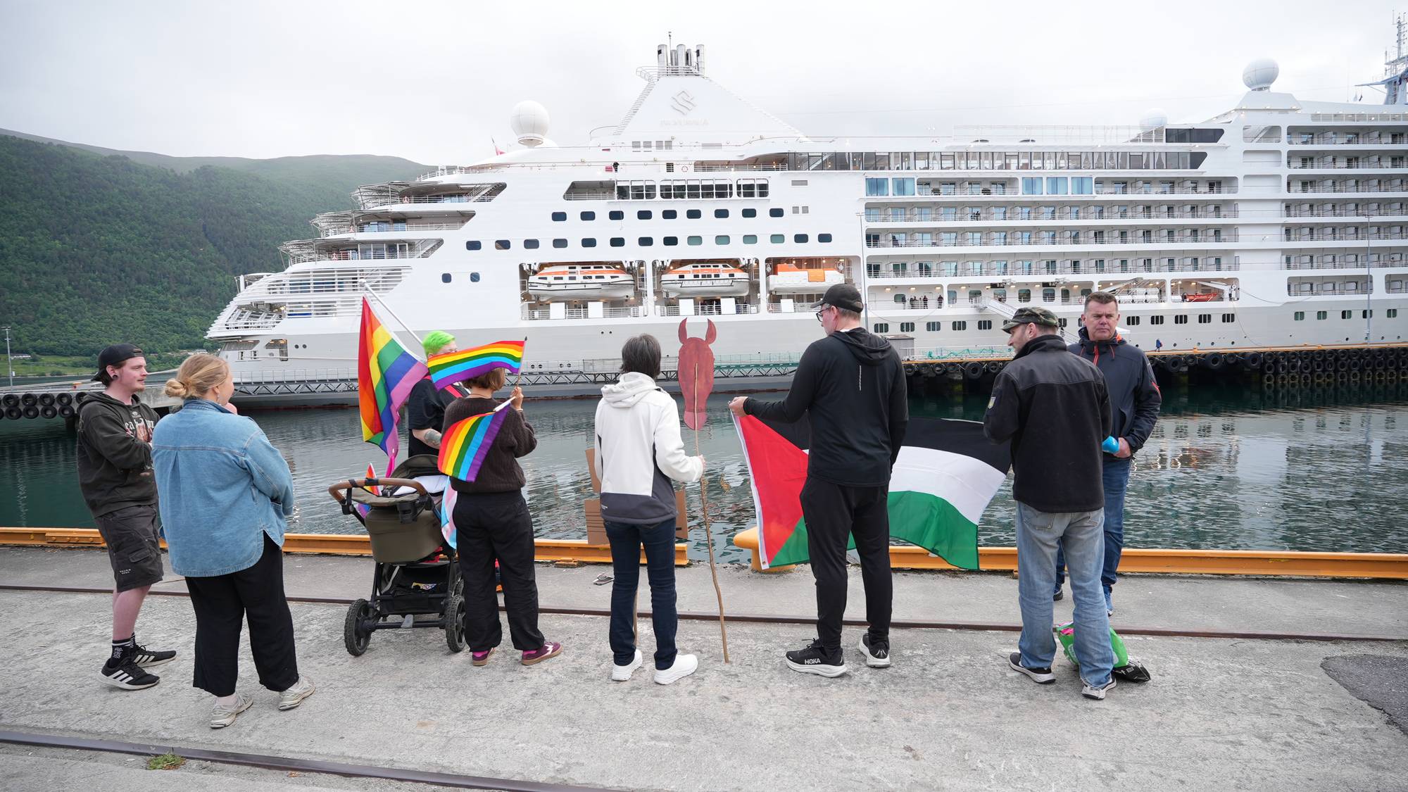 En gruppe mennesker står samlet ved en brygge. Noen holder regnbueflagg, mens andre holder et palestinsk flagg. En stor hvit cruiseskip ligger til kai bak dem. (Bildebeskrivelsen er laget av en KI-tjeneste)