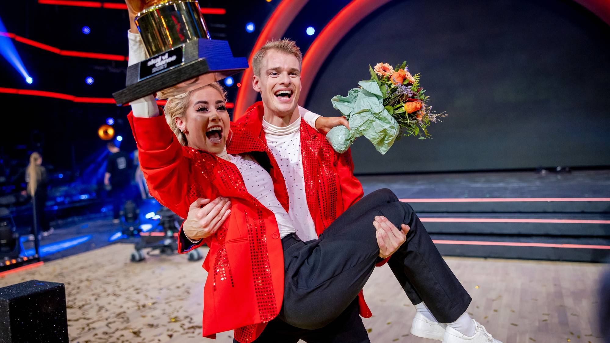 En mann bærer en kvinne som holder et trofé og en blomsterbukett. Begge er kledd i glitrende røde jakker og smiler. Scenen er dekket av konfetti. (Bildebeskrivelsen er laget av en KI-tjeneste)
