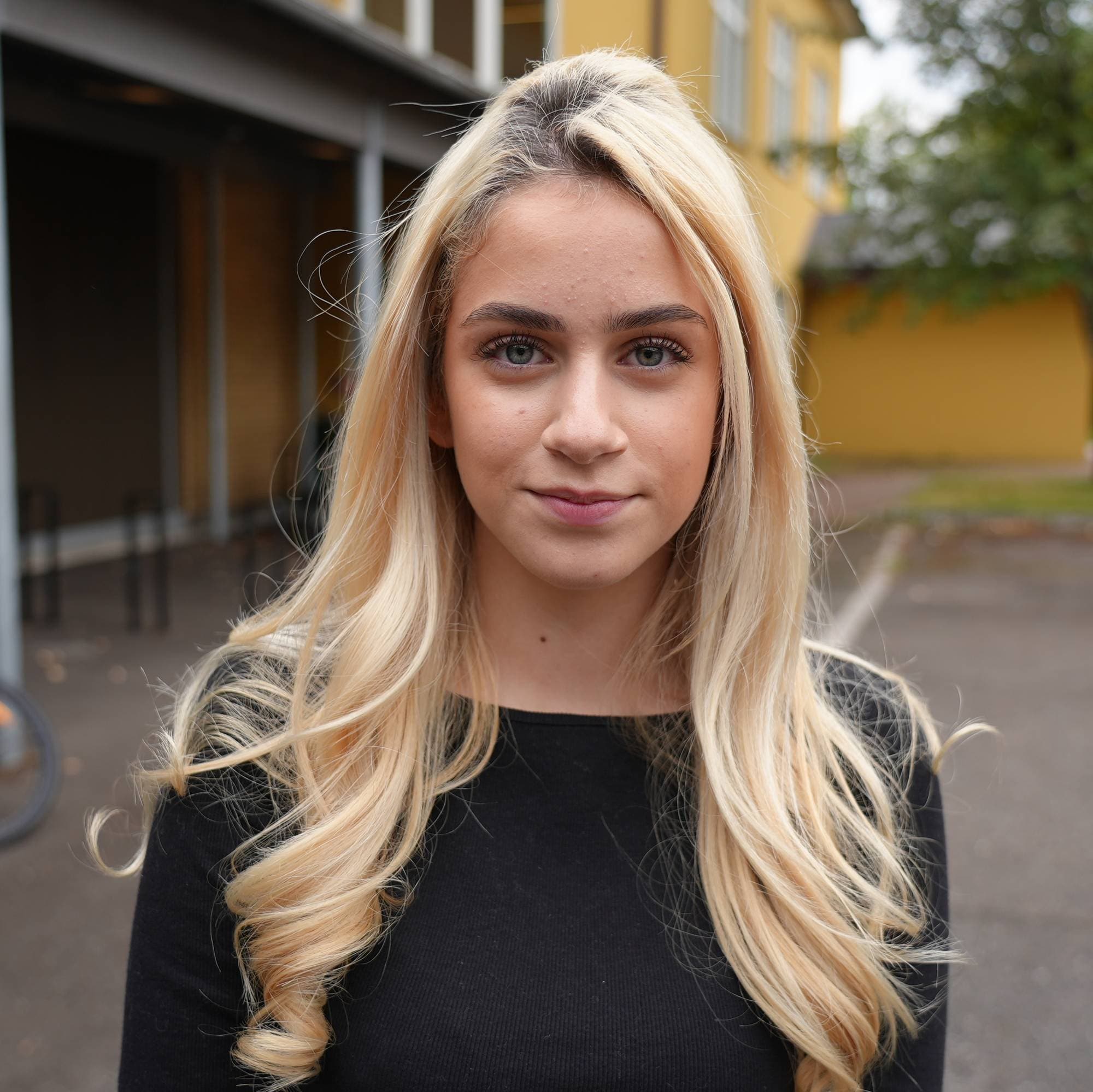 Portrett av en ung kvinne med langt blondt hår foran en skolebygning.