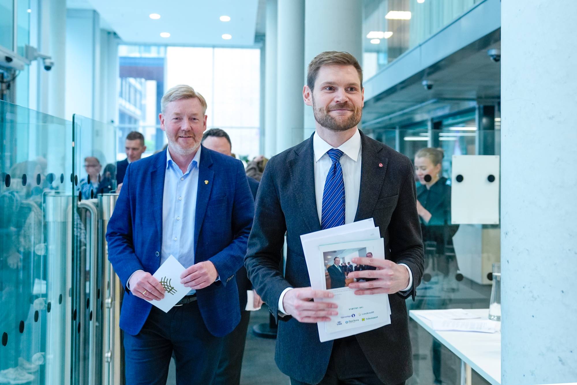 To menn går mot kamera med papirer i hendene. Den ene mannen til venstre har blå dress og lys skjorte. Mannen til høyre har mørk dress, blå slips og holder flere ark. De er i en lys bygningskorridor. (Bildebeskrivelsen er laget av en KI-tjeneste)
