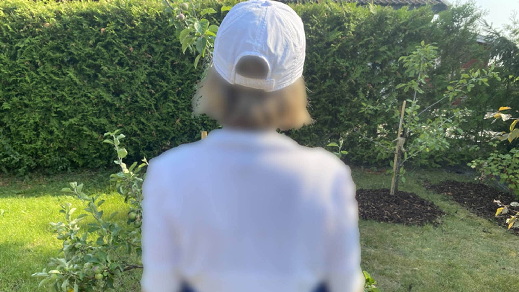 Bildet viser en anonymisert kvinne bakfra. Hun står i en hage og har på seg en hvit caps og en hvit T-skjorte.