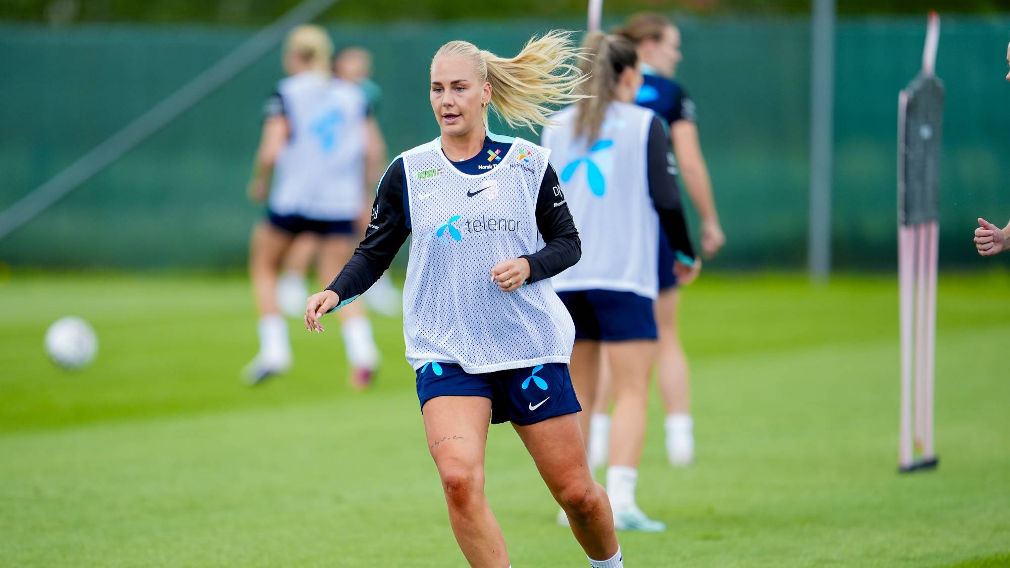En fotballspiller i blå drakt og hvit vest trener på en gressbane. Flere spillere i bakgrunnen trener også. De er på en fotballbane med grønt gress. (Bildebeskrivelsen er laget av en KI-tjeneste)