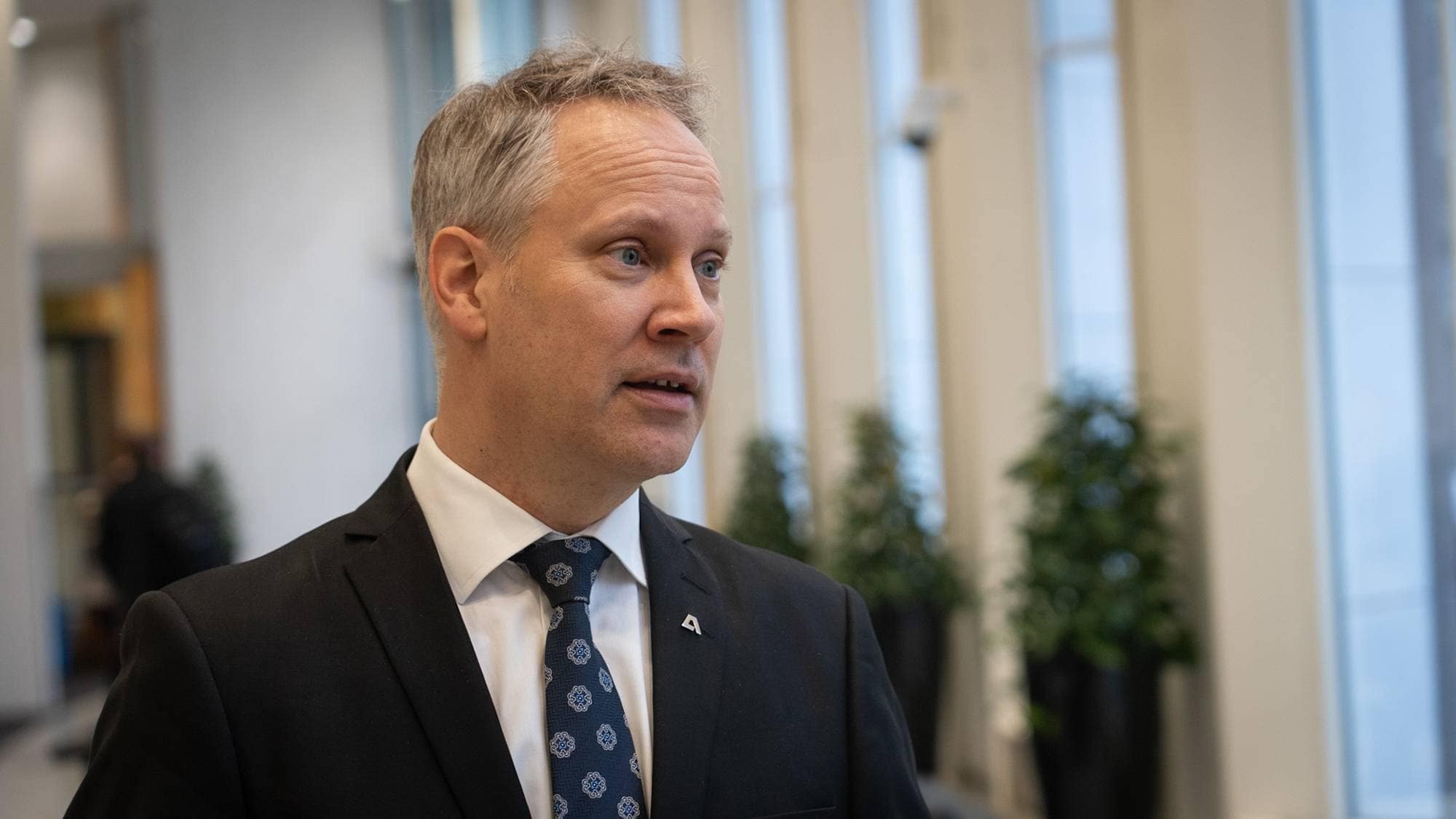 En mann iført dress og slips står i et lysfylt rom med store vinduer. I bakgrunnen er det grønne planter. Mannen ser mot venstre og virker fokusert. Omgivelsene gir et moderne og profesjonelt inntrykk. (Bildebeskrivelsen er laget av en KI-tjeneste)