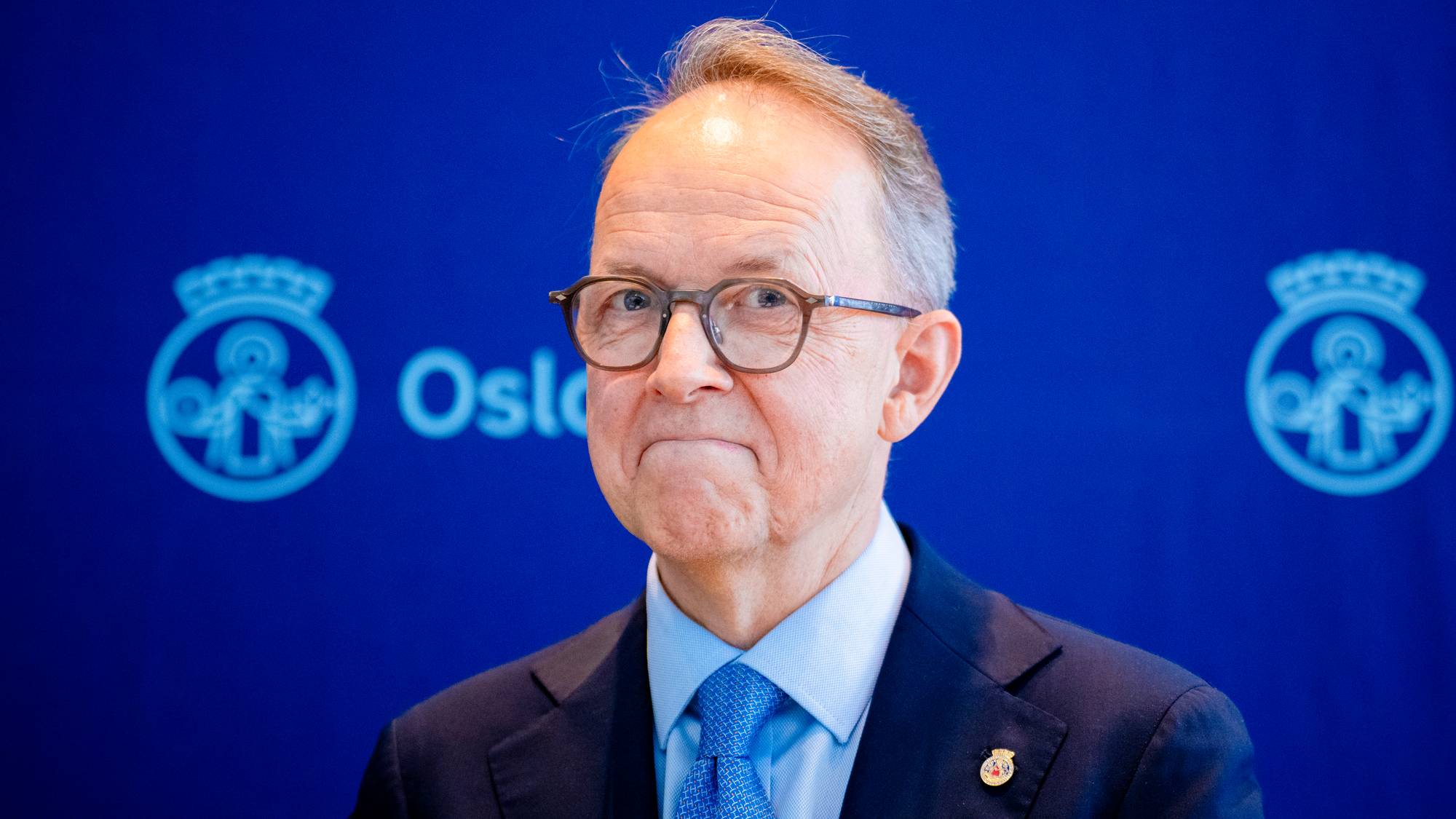En eldre mann med briller ser mot kameraet. Han er kledd i mørk dress, lyseblå skjorte og blått slips. Bak ham er en mørkeblå bakgrunn med logoer. (Bildebeskrivelsen er laget av en KI-tjeneste)
