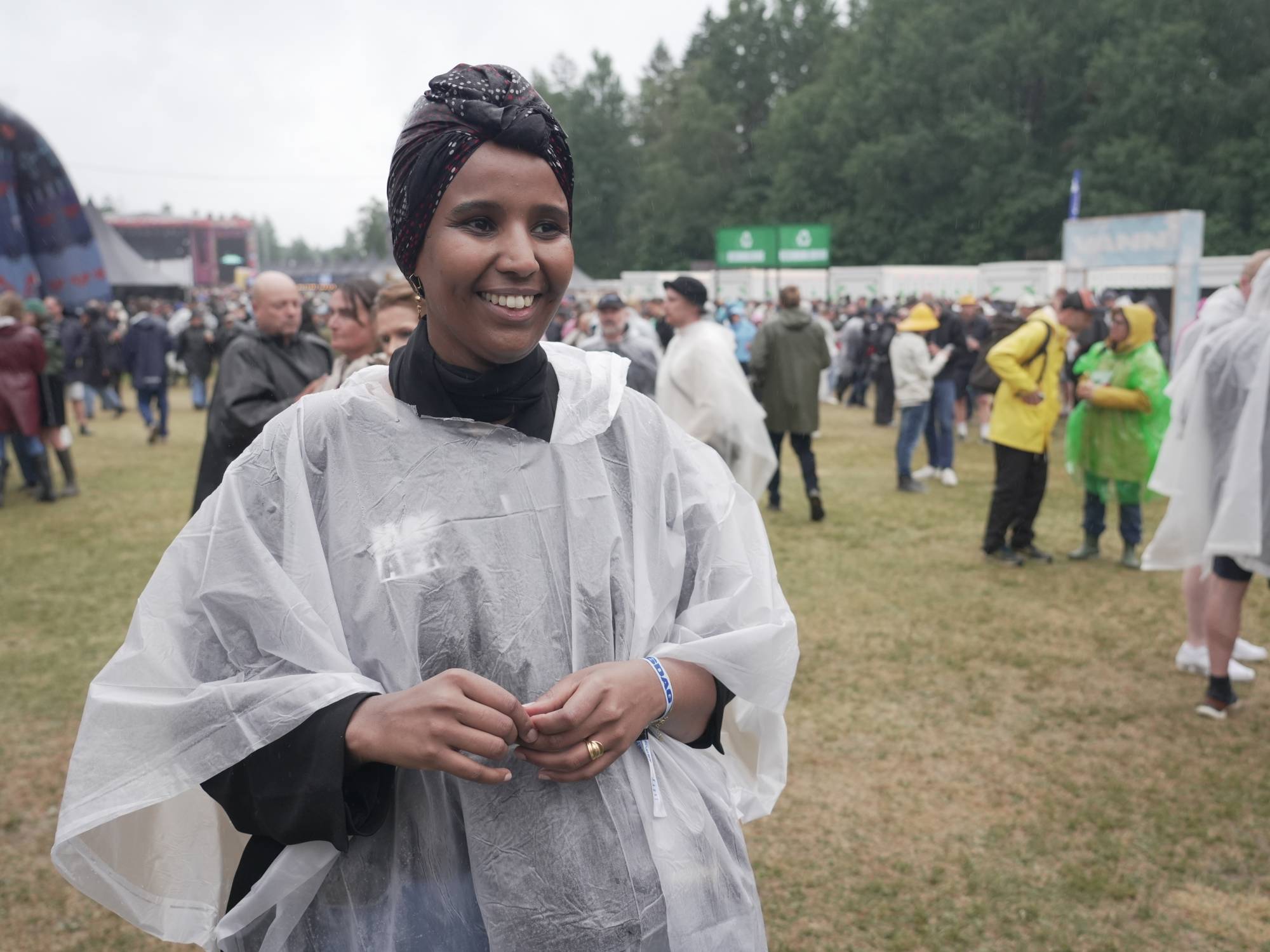 Smilende kvinne i regnponcho på festivalområde med publikum i bakgrunnen. (Bildebeskrivelsen er laget av en KI-tjeneste)