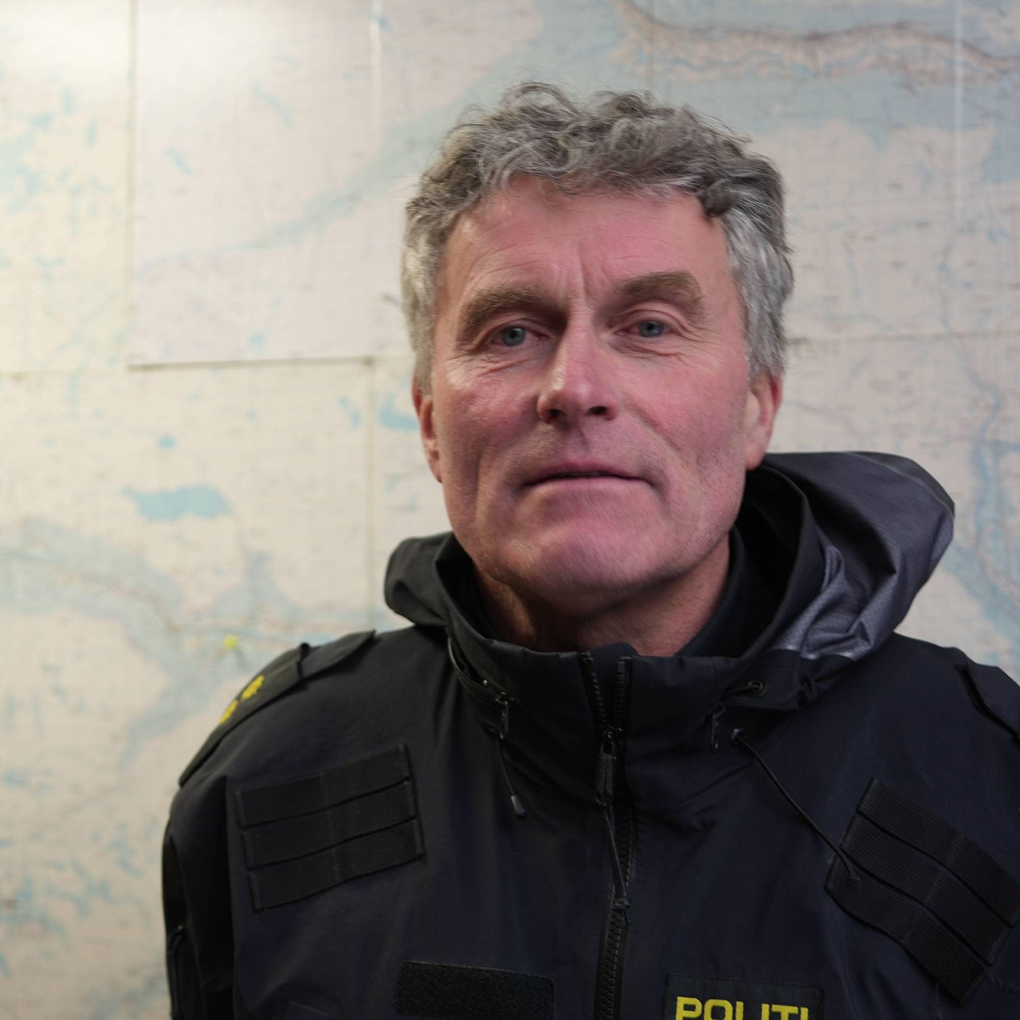 En politimann i uniform står foran et kart. Han har grått hår og ser mot kameraet. Jakken hans er mørkeblå med gule bokstaver som sier "POLITI". Kartet bak ham dekker en stor del av veggen. (Bildebeskrivelsen er laget av en KI-tjeneste)