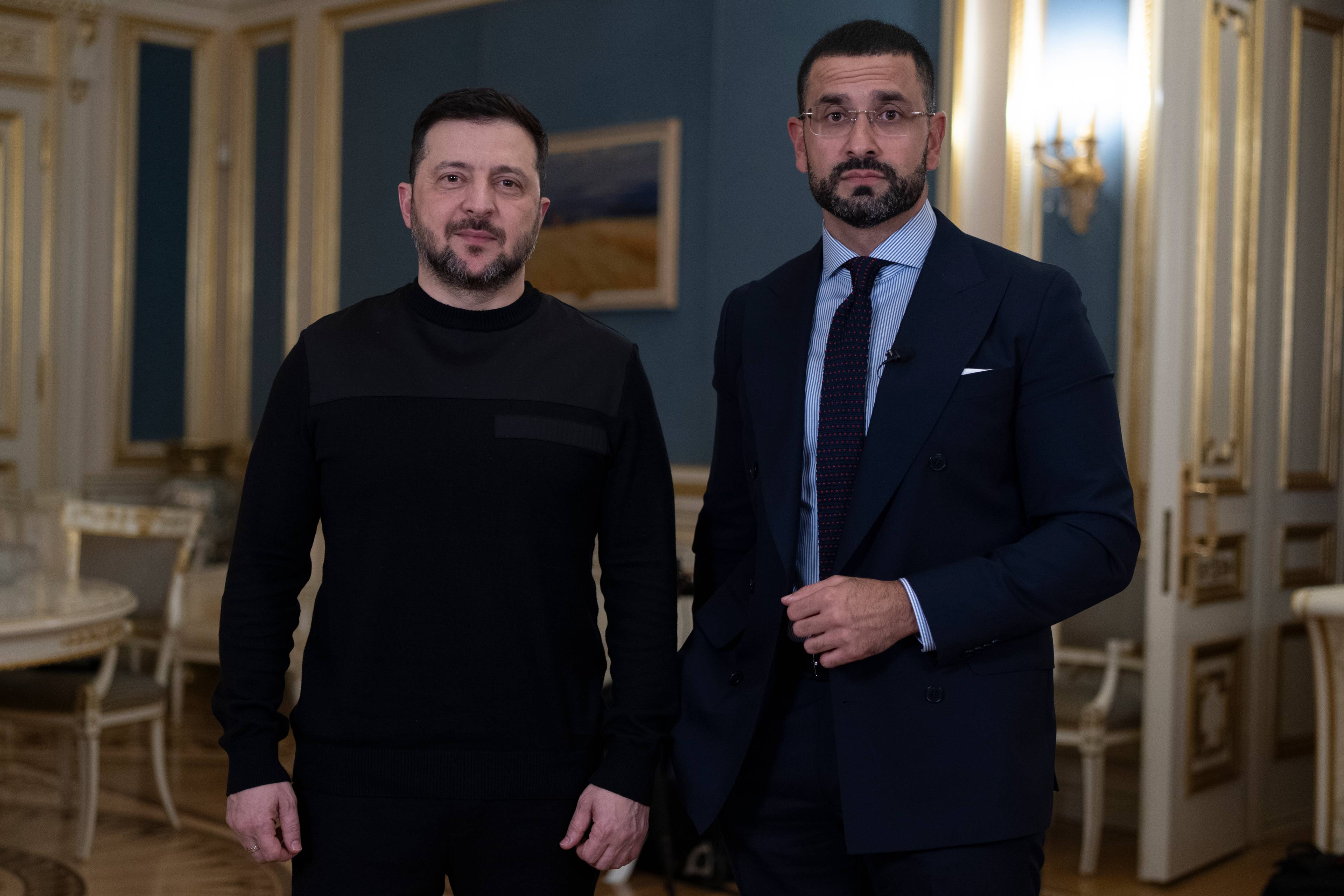 Ukrainas president Volodymyr Zelenskyj står ved siden av en mann i mørk dress.Begge mennene ser mot kamera.Rommet er overdådig dekorert med gulldetaljer og et maleri på veggen.Mennene er fotografert i et intervju. (Bildebeskrivelsen er laget av en KI-tjeneste)