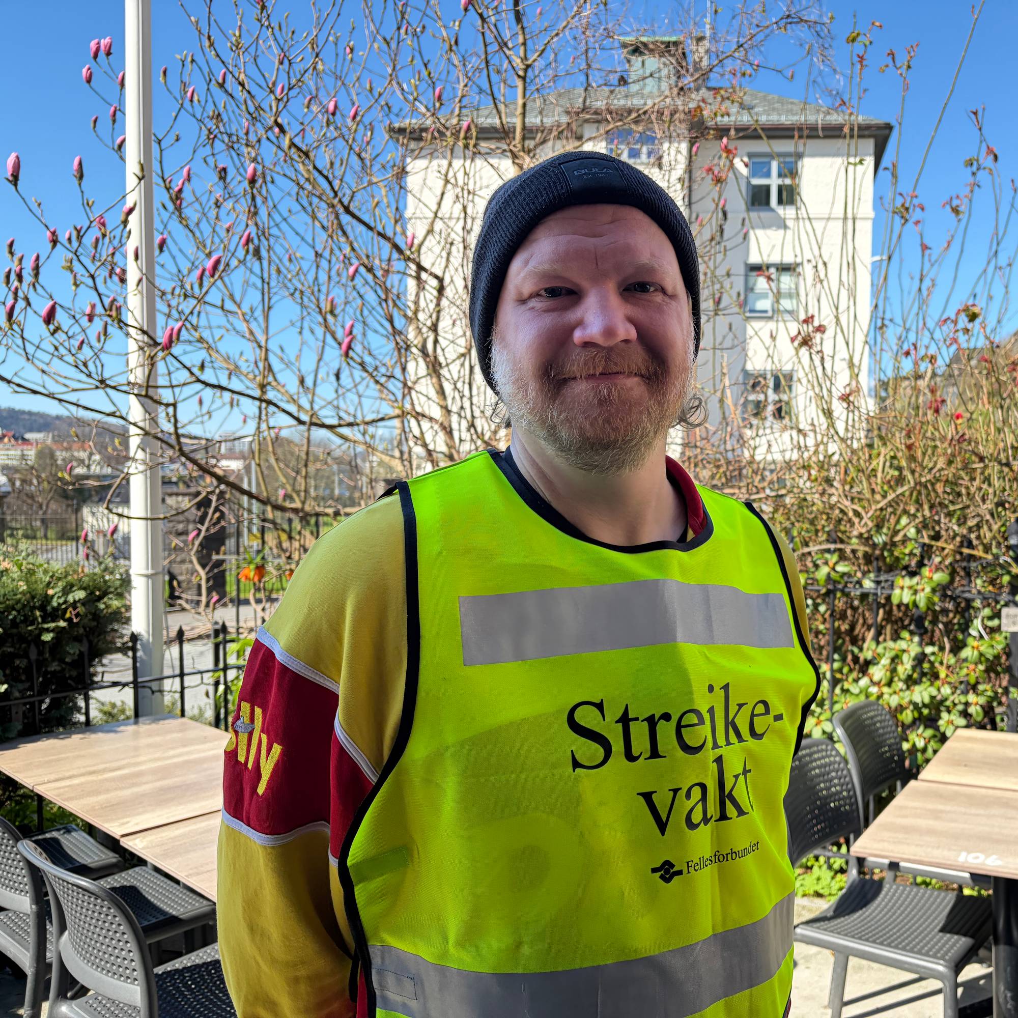 En mann med svart lue og gul vest med teksten "Streikevakt" står foran en bygning. Mannen smiler mot kamera. Bak ham er det trær, bord og stoler. 